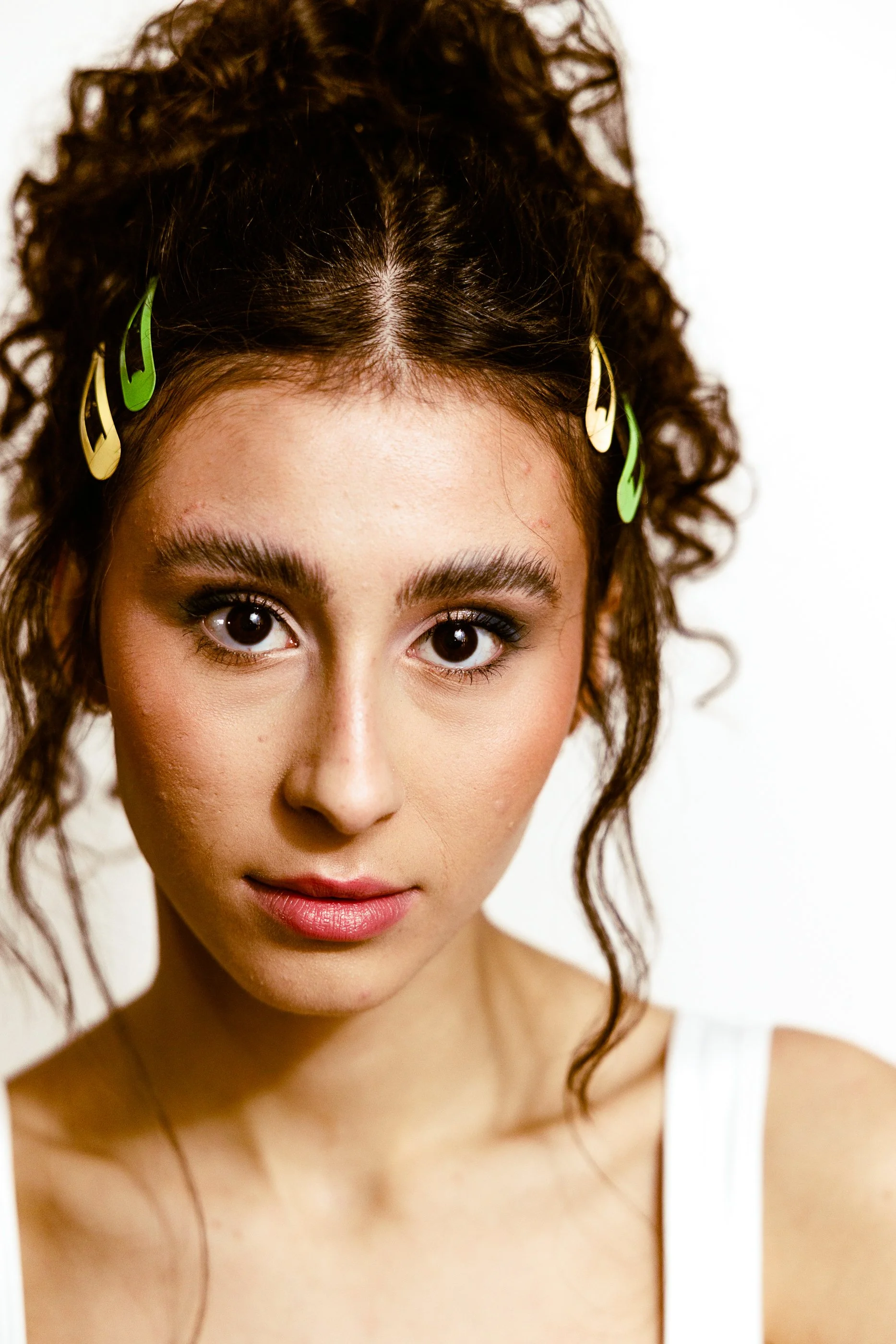 Close-up of a young woman with curly brown hair held back with colorful hair clips, wearing a white top, looking directly at the camera at the hair salon in Jersey City.