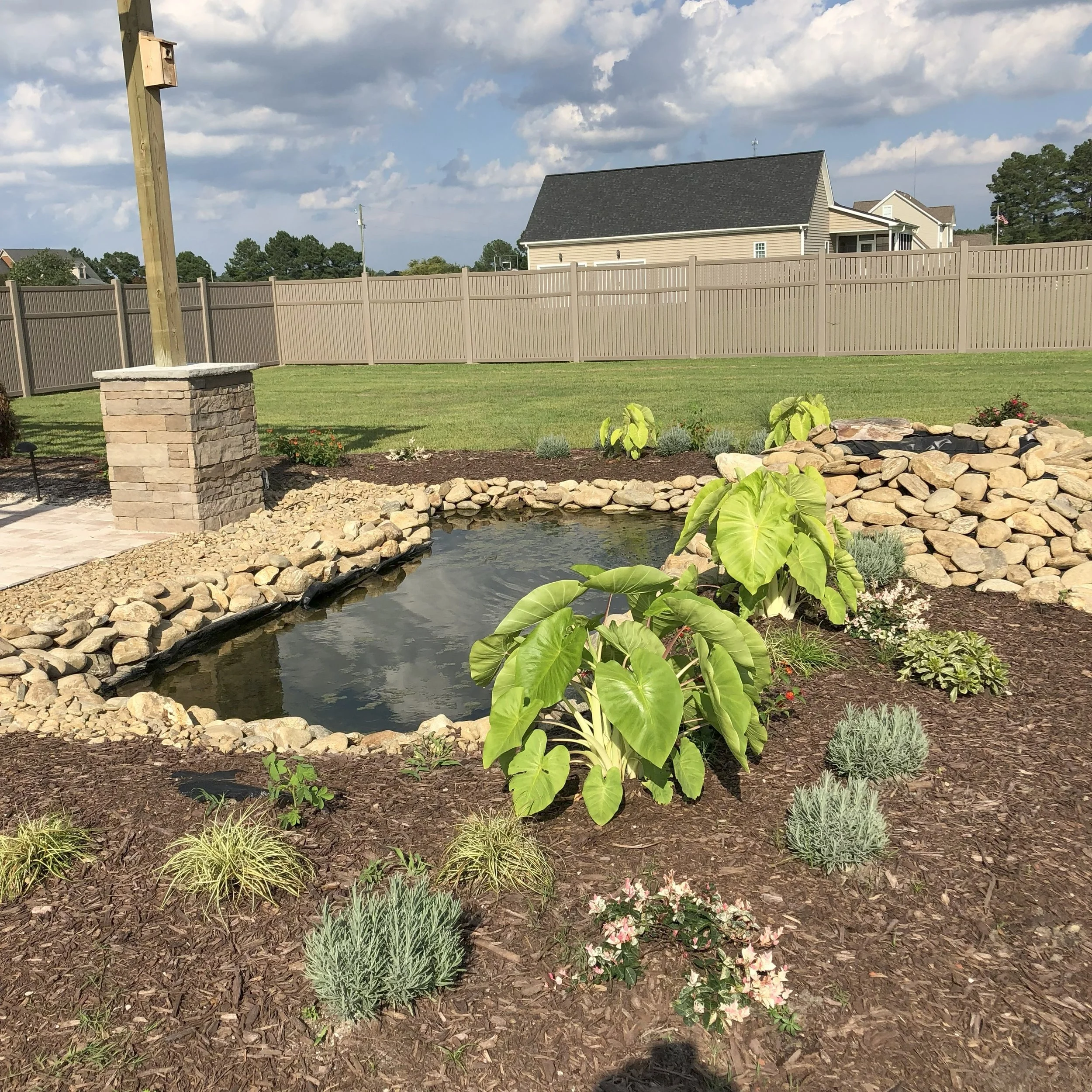 A garden with a small pond surrounded by rocks, various green plants, a stone pillar, and a wooden fence in the background under a cloudy sky.
