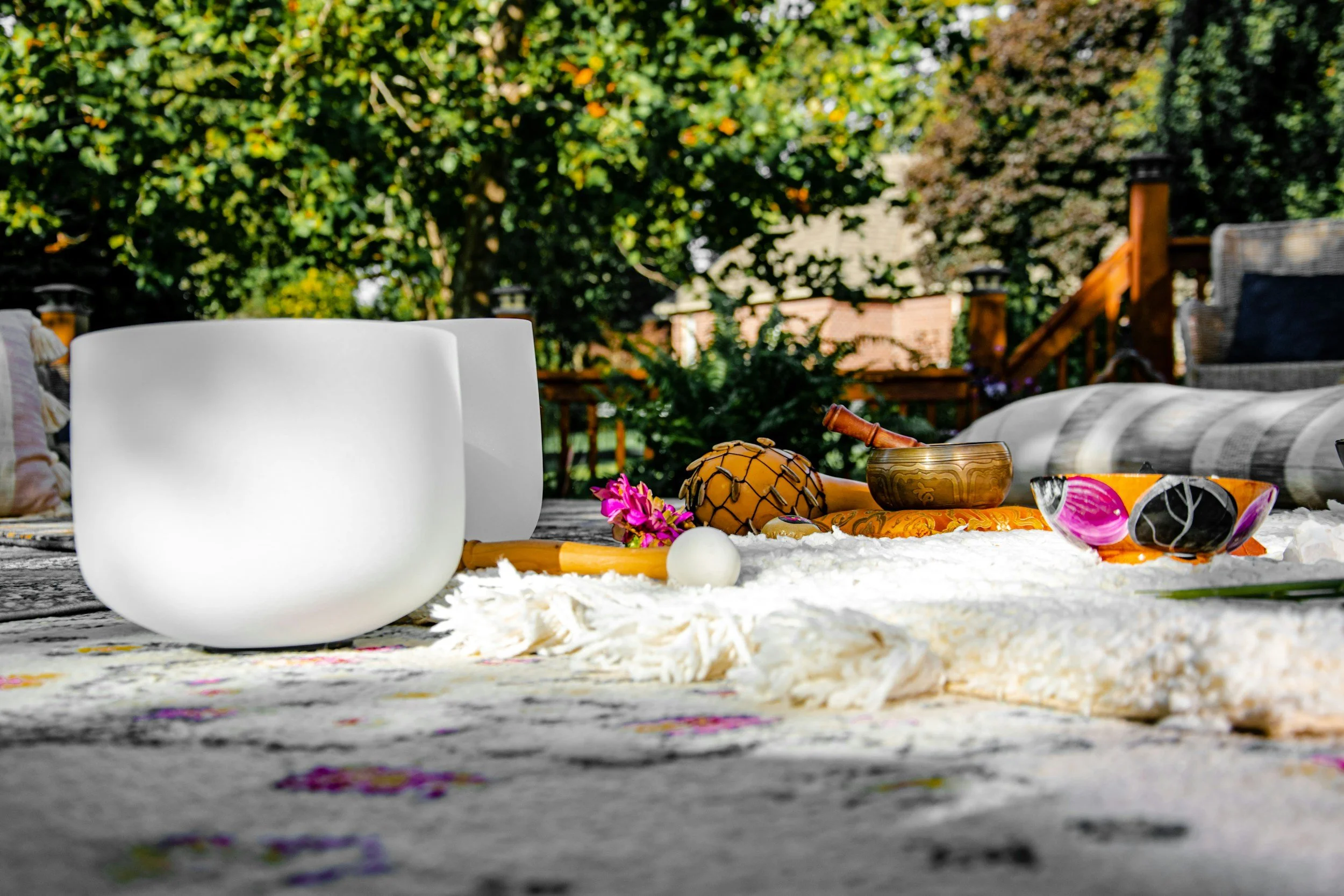 A backyard outdoor space with musical instruments, such as a crystal singing bowl and a wooden bowl, placed on a white textured blanket. There is a small pink flower, a white ball, and a carved wooden object near the instruments. The background features a wooden deck railing, trees, and a house.