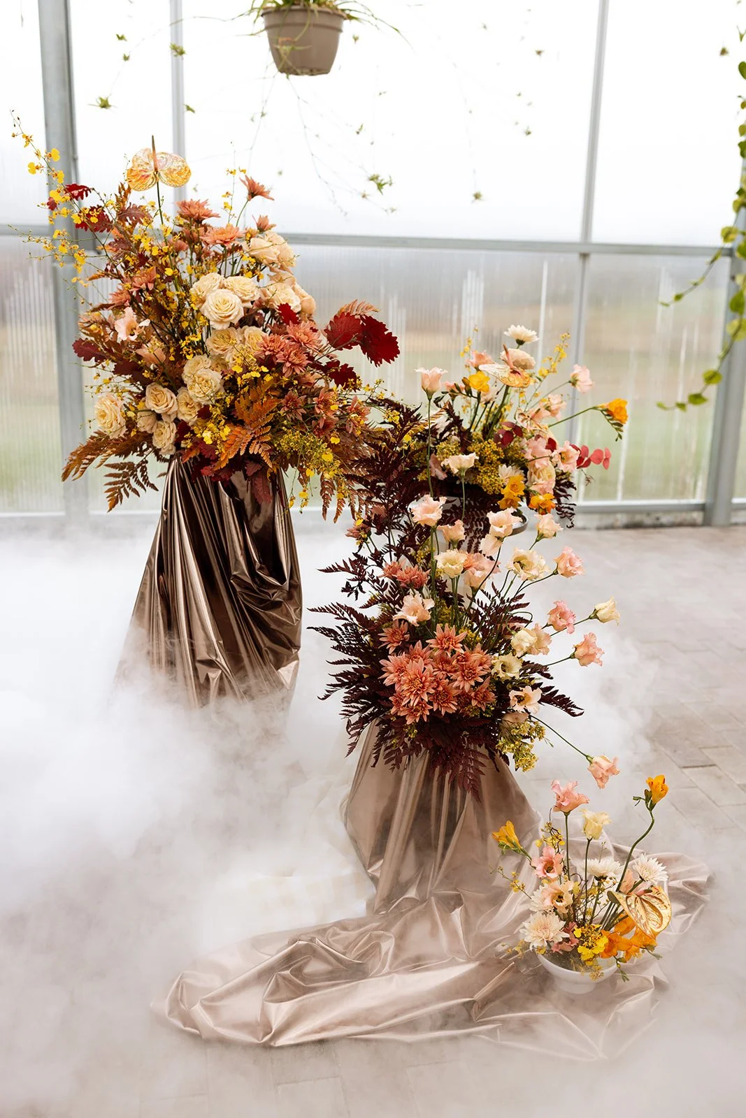 Decorative floral arrangements with roses, chrysanthemums, and other flowers in metallic vases and bowls, with draped fabric and mist or fog on the floor in a greenhouse setting.