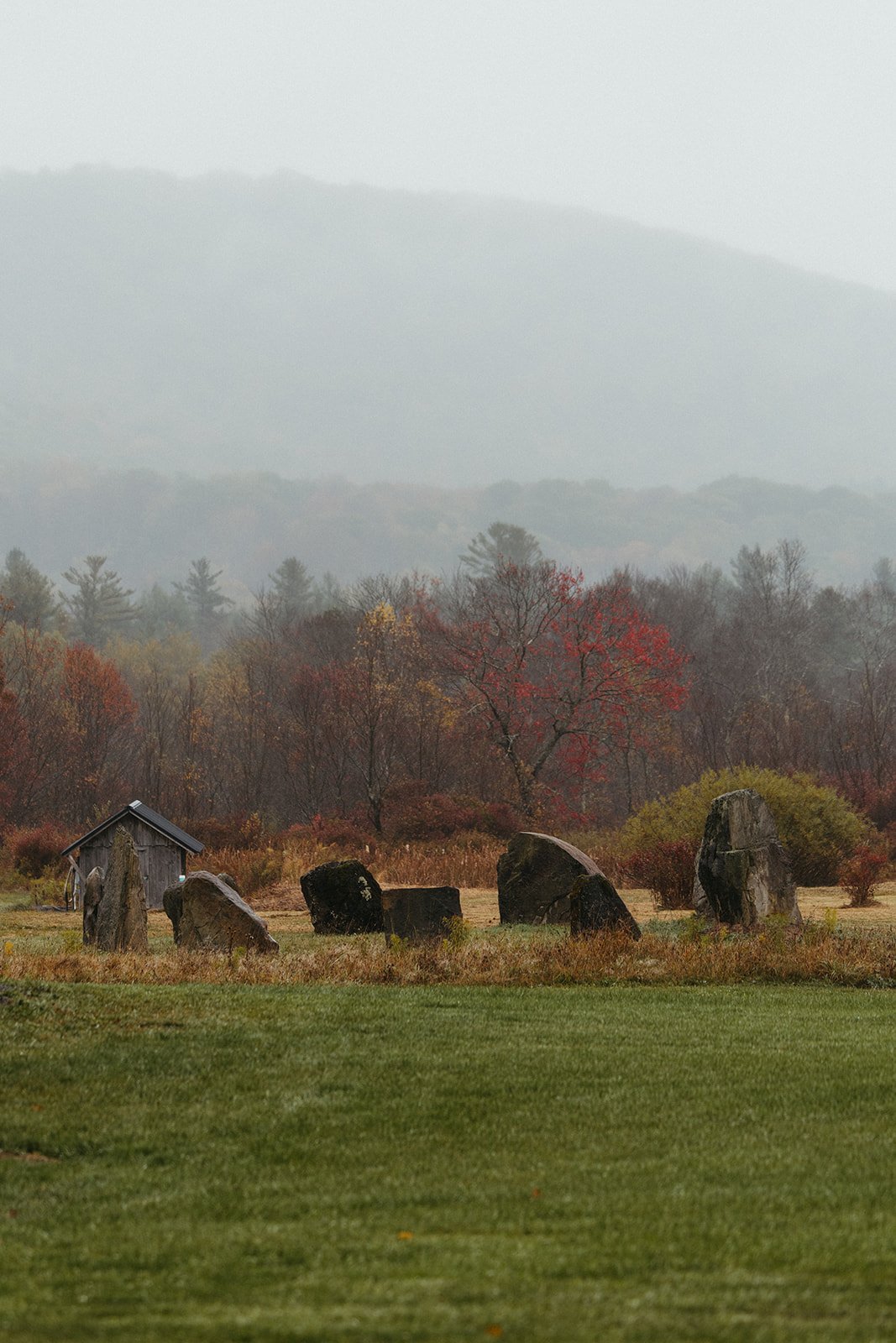 A landscape with a grassy foreground, a circle of large stones, a small wooden shed, and colorful trees with fall foliage, with mountains and fog in the background.