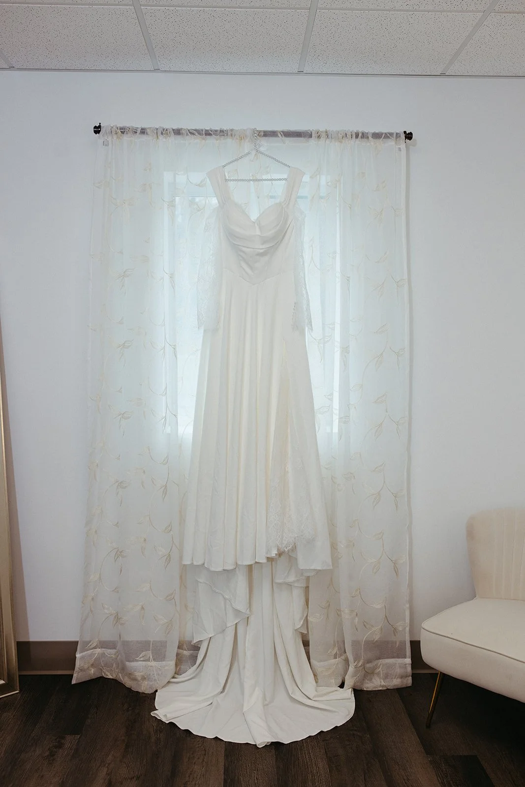 White wedding dress hanging on a hanger in front of a window with sheer curtains.