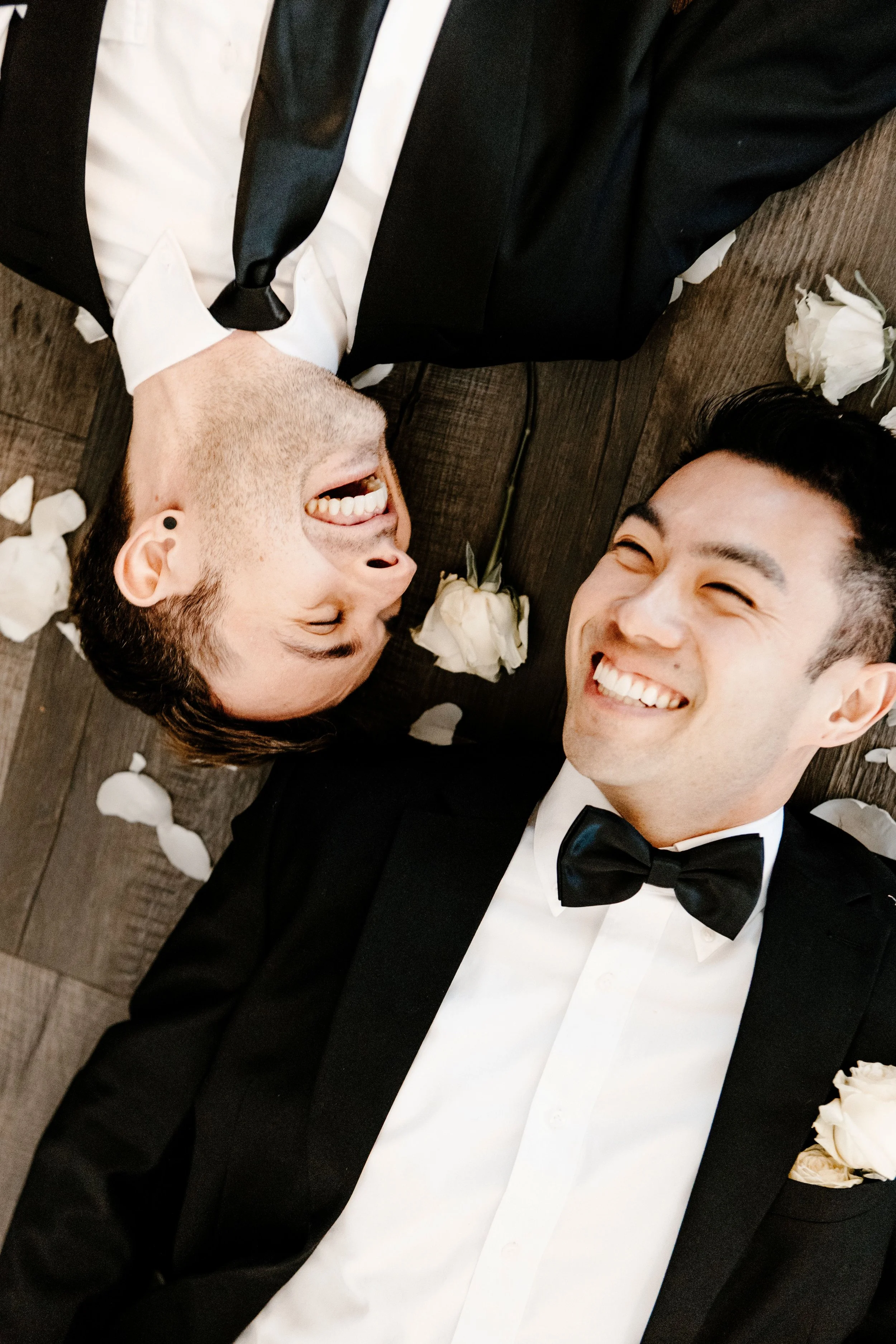 Two men in tuxedos lying on a wooden floor surrounded by white rose petals, laughing and smiling at each other.