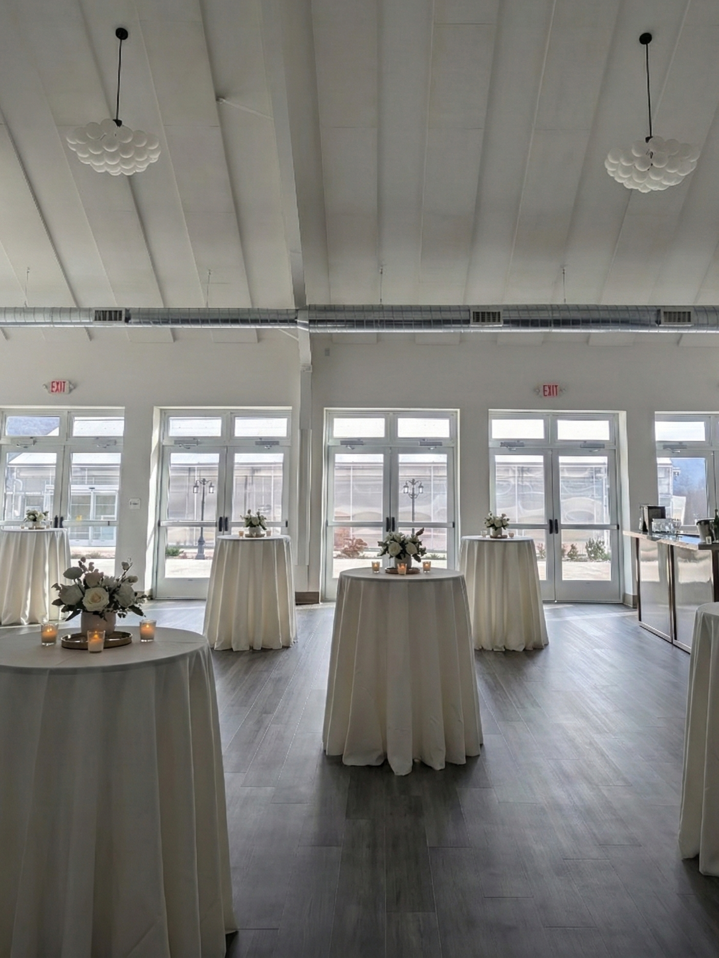 Indoor event space decorated with high-top tables covered with white tablecloths, decorated with floral centerpieces and candles, large windows and glass doors in the background, modern ceiling lights, and wood flooring.