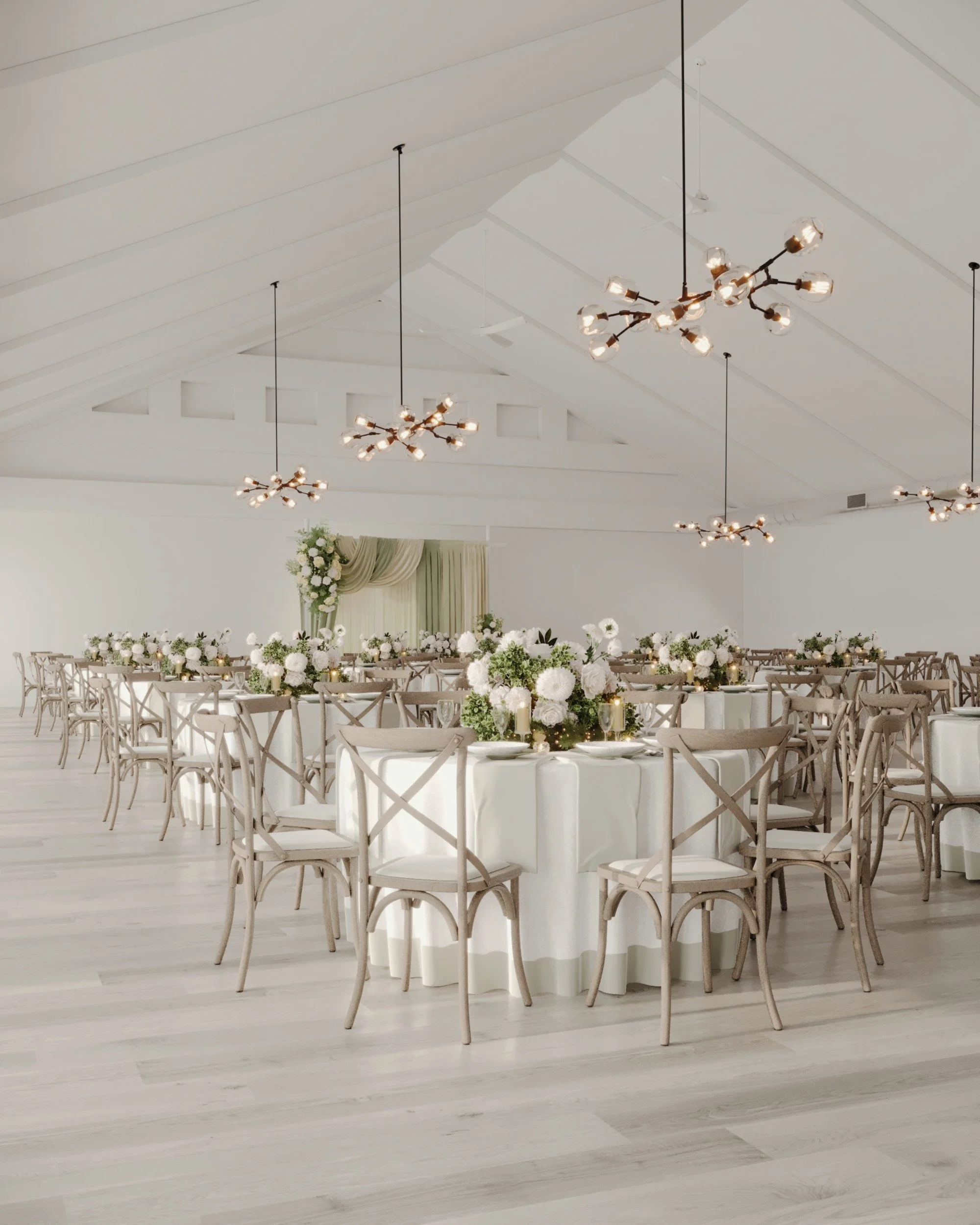 A spacious, well-lit event hall decorated for a wedding reception with round tables covered in white tablecloths, each with floral centerpieces featuring white and green flowers, surrounded by wooden chairs with cross-back design, and elegant pendant lights hanging from a high ceiling.
