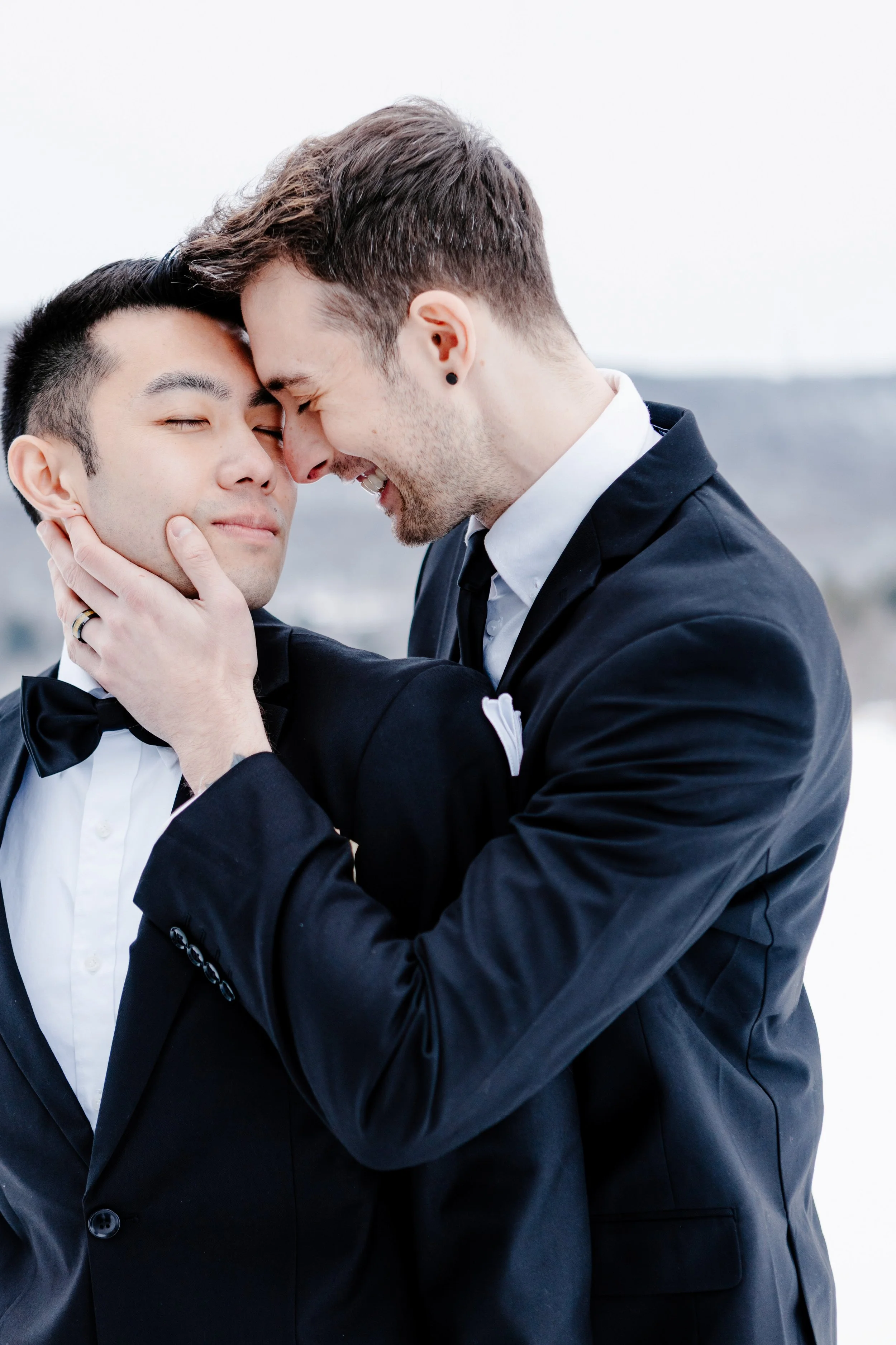 Two men in formal suits sharing an intimate moment, pressing foreheads together outdoors against a cloudy sky.