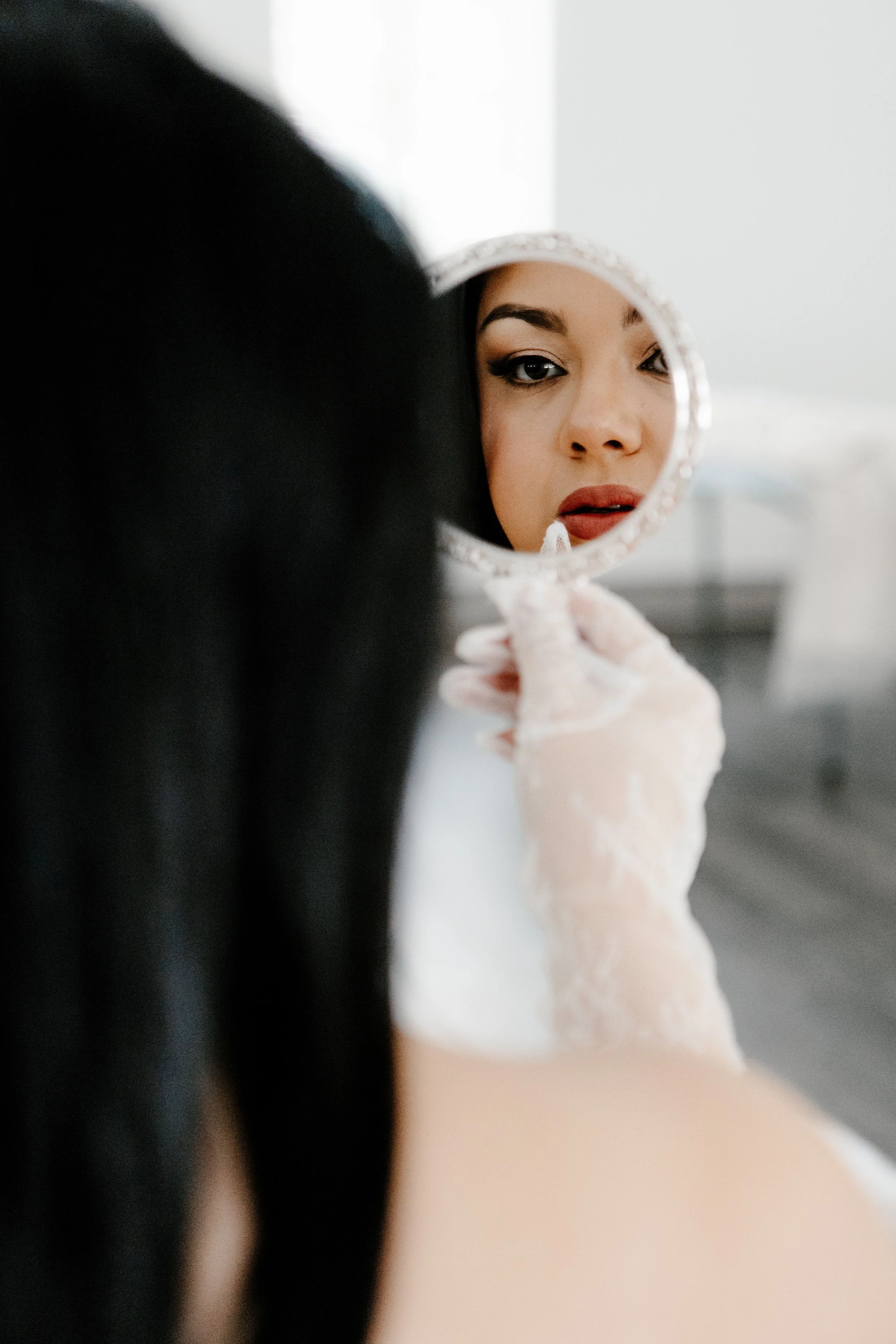 A woman with makeup looking at her reflection in a round hand mirror.