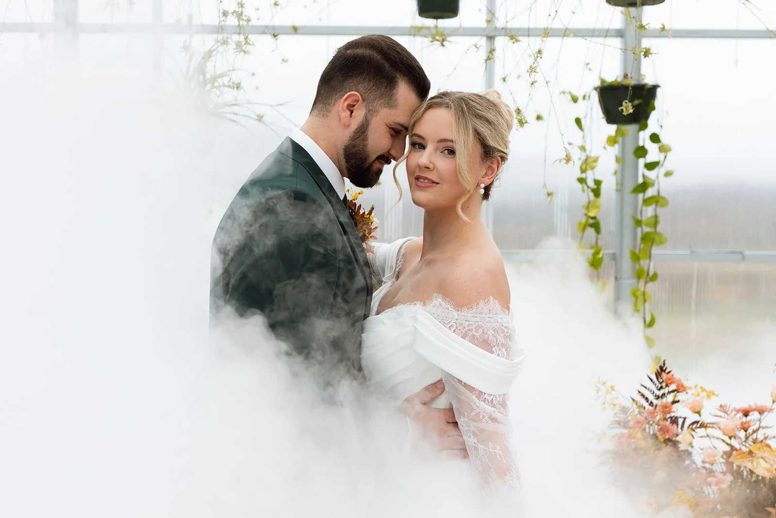 A bride and groom pose closely together in a greenhouse, surrounded by mist and hanging plants, with flowers in the background.