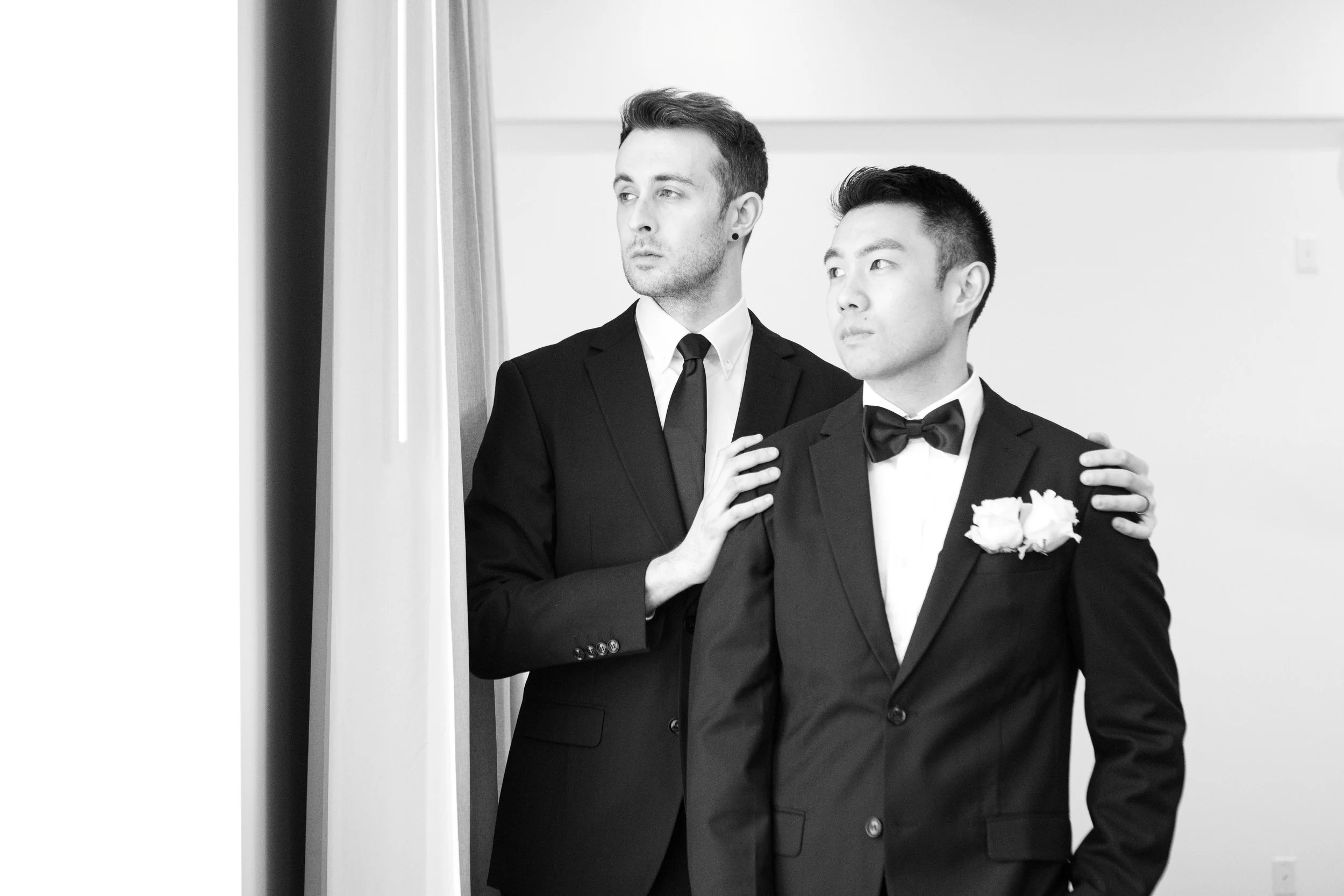 Two men in tuxedos standing indoors, one with a flower boutonniere, looking off to the side, with one man resting his hand on the other's shoulder.