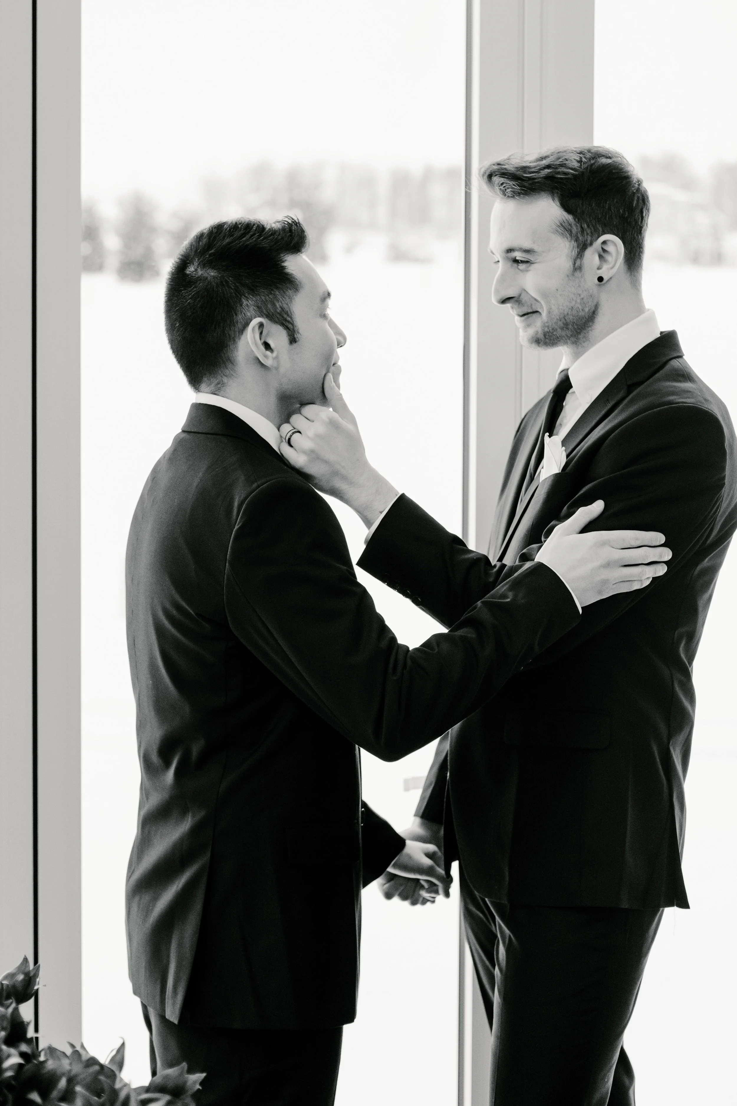 Two men in suits holding hands and touching faces in an intimate moment indoors with a large window behind them.