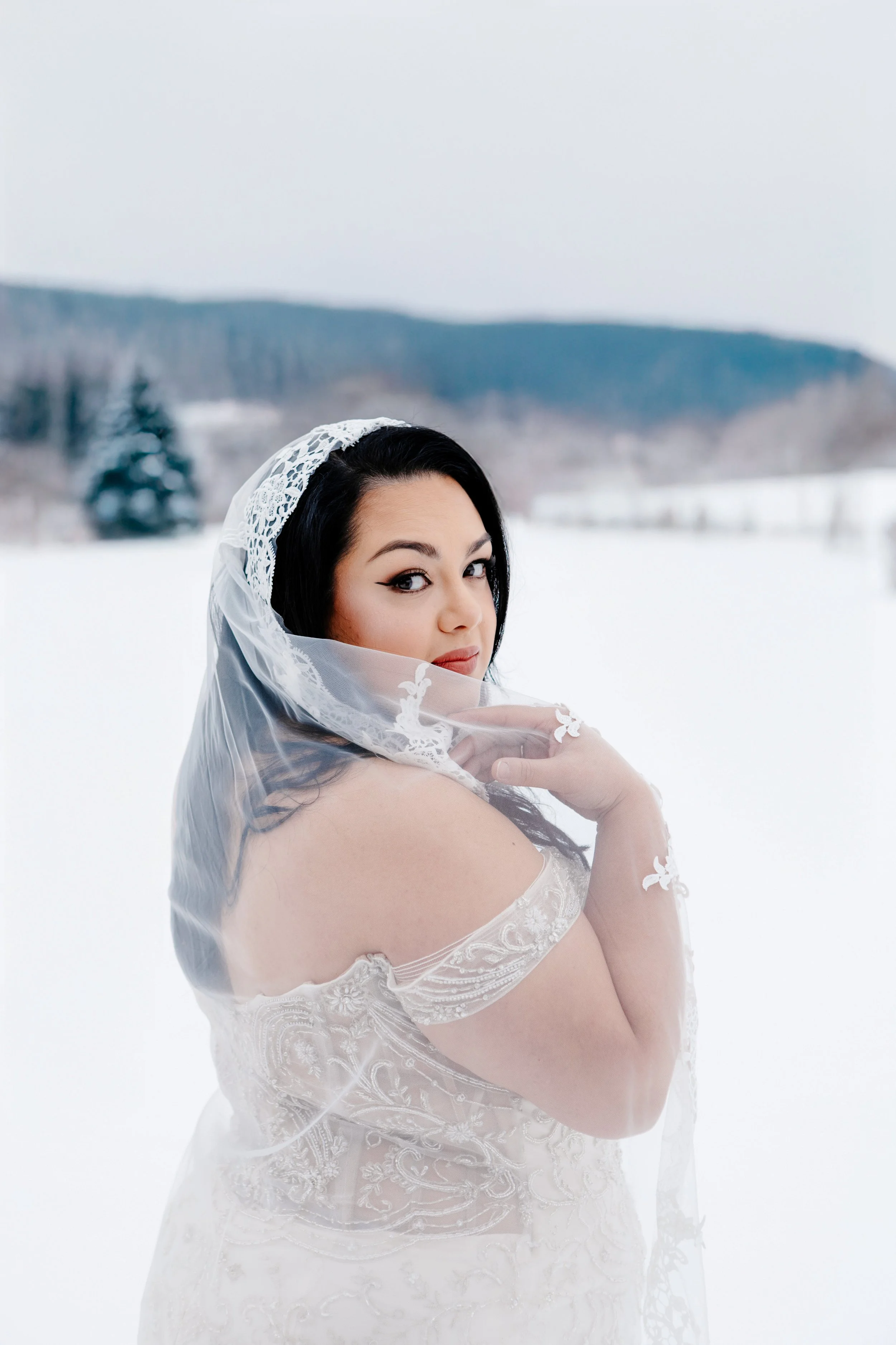 A woman in a white lace wedding dress standing outside in a snowy landscape, looking over her shoulder at the camera with a veil draped over her head.
