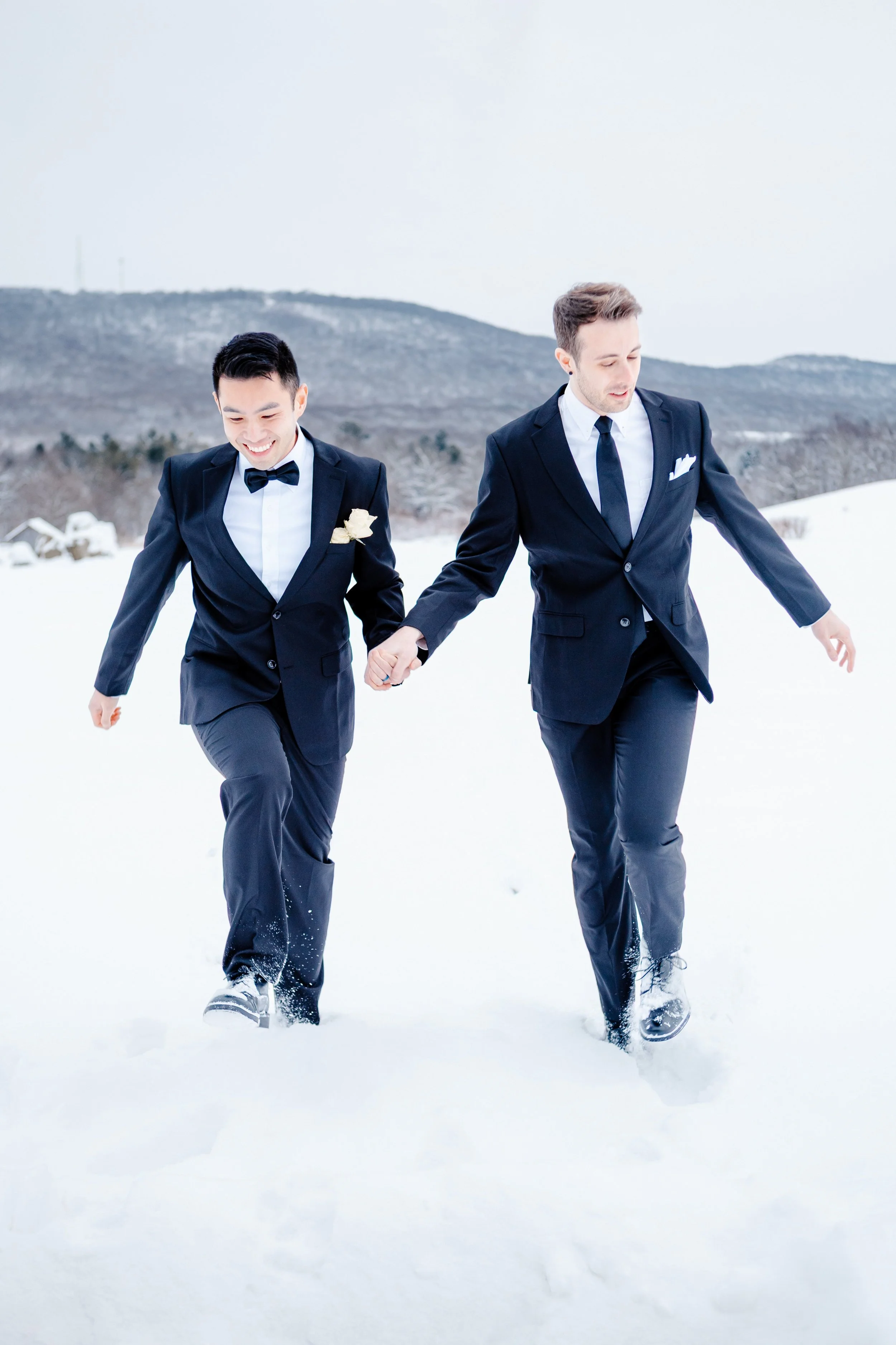 Two men in tuxedos walking hand in hand in snow, smiling, with a mountain landscape in the background.