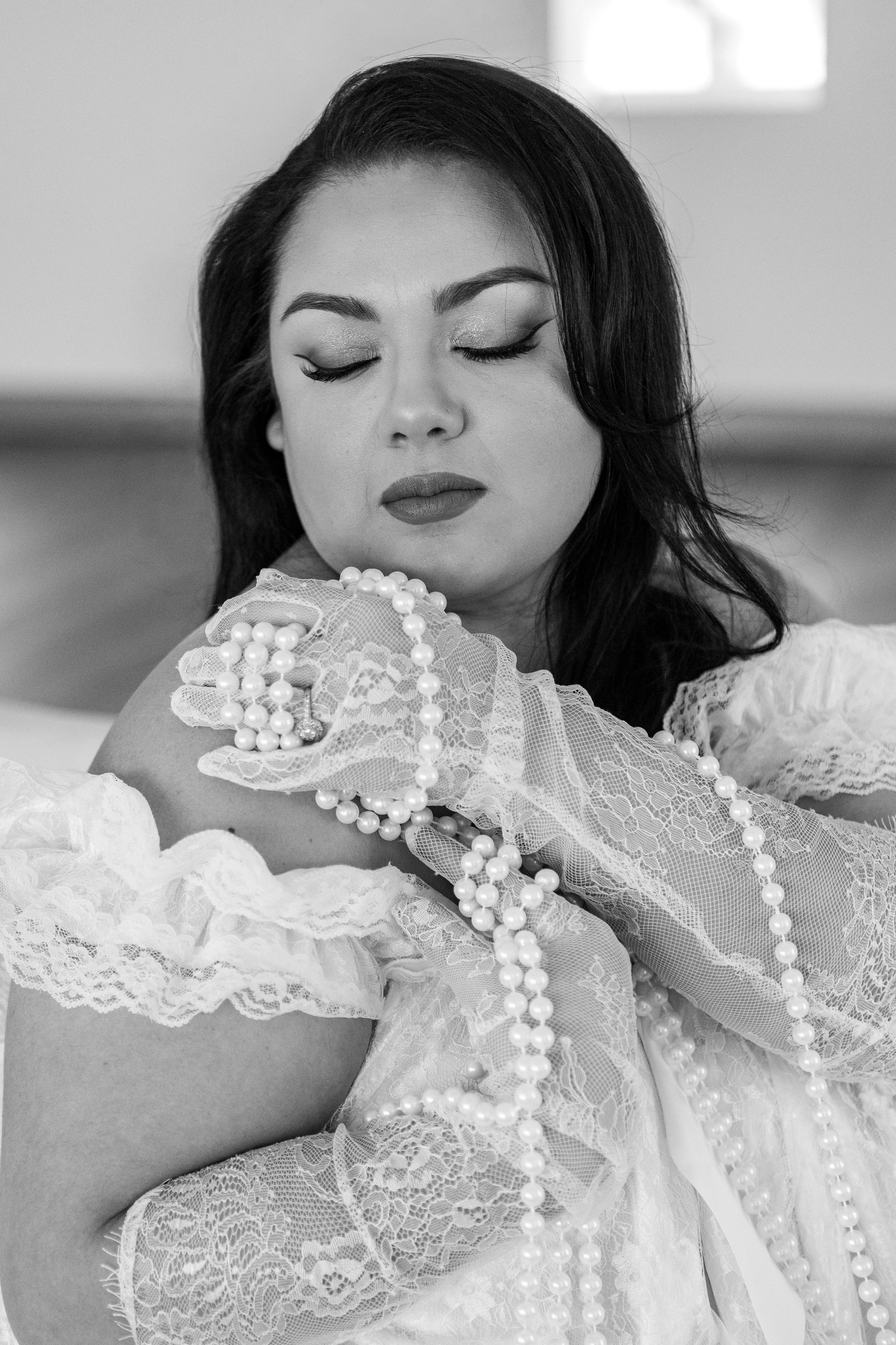 A woman with dark hair and makeup, dressed in lace gloves and holding pearl necklaces, with her eyes closed.