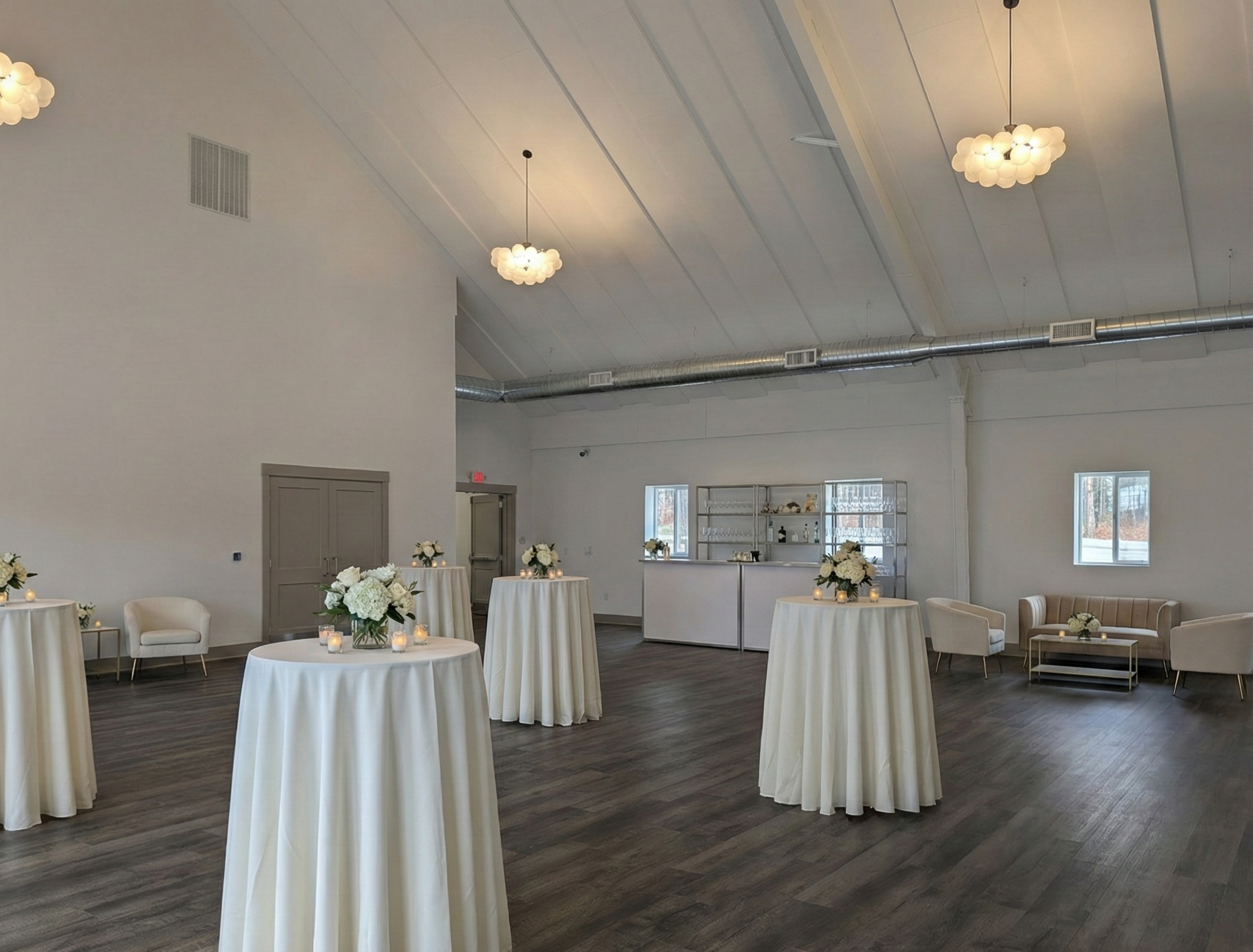 An elegant event space with high white ceiling, wooden flooring, and soft lighting. Five round tables with white tablecloths are decorated with flower arrangements and candles. Comfortable chairs and a sofa seating area are along the walls, and a bar in the background.