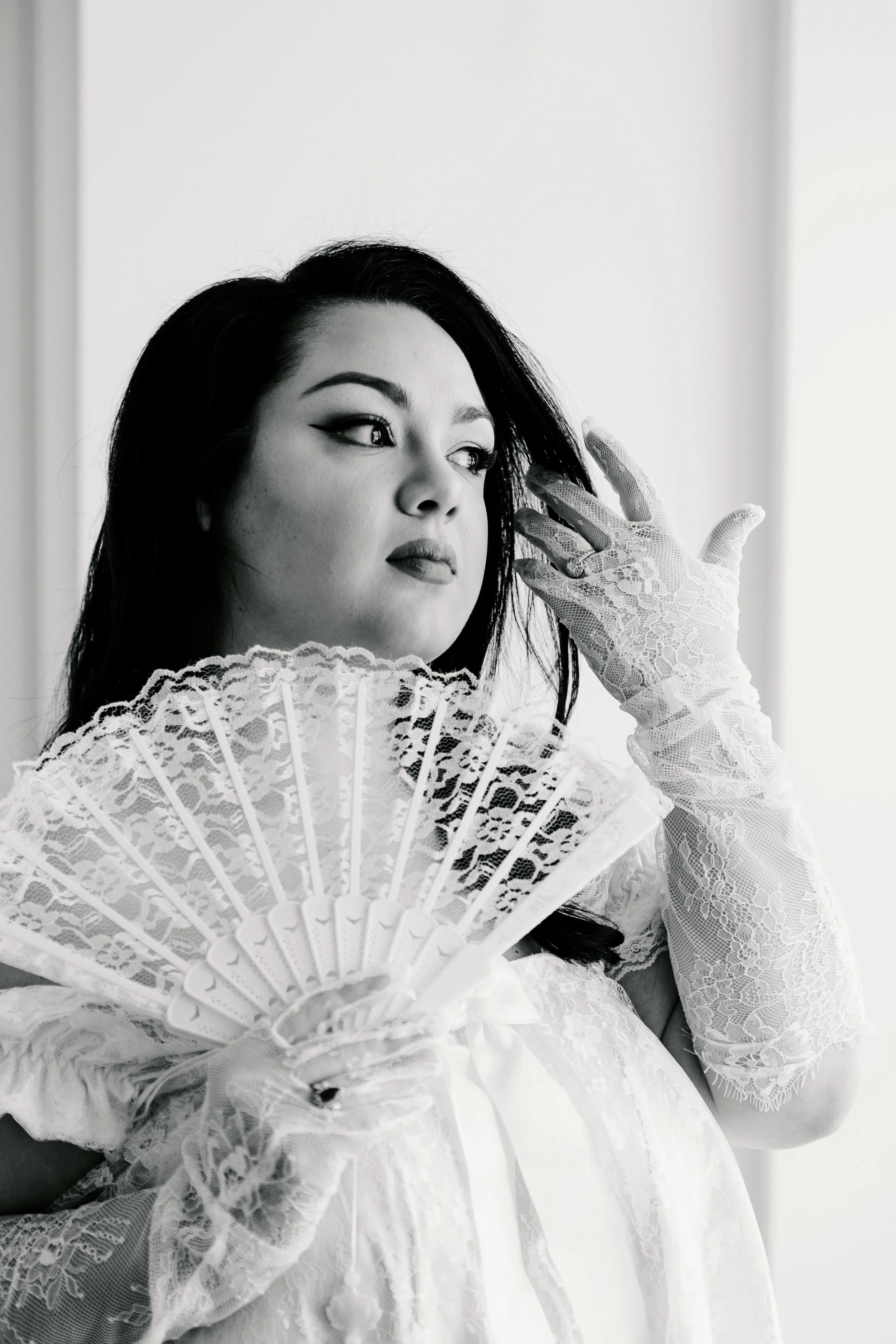 A woman with dark hair, dressed in vintage lace gloves and a dress, holds an ornate lace fan near her chest. She is looking to the side with a contemplative expression.