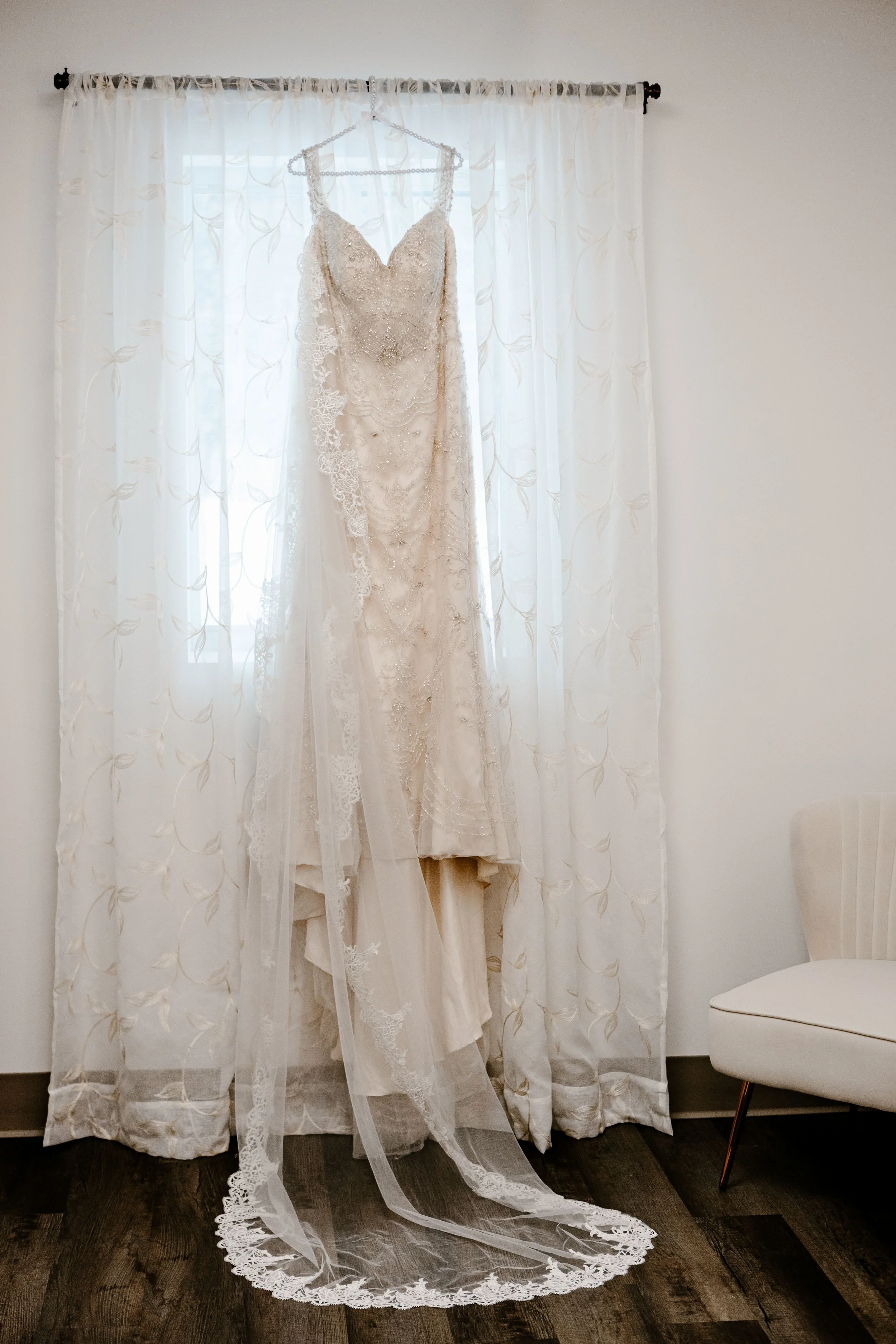 A wedding dress hanging on a hanger in front of white curtains.