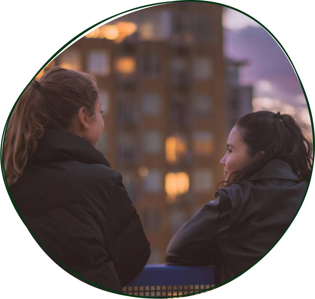Two women are sitting on a bench, talking and smiling at each other, with a cityscape and sunset in the background.