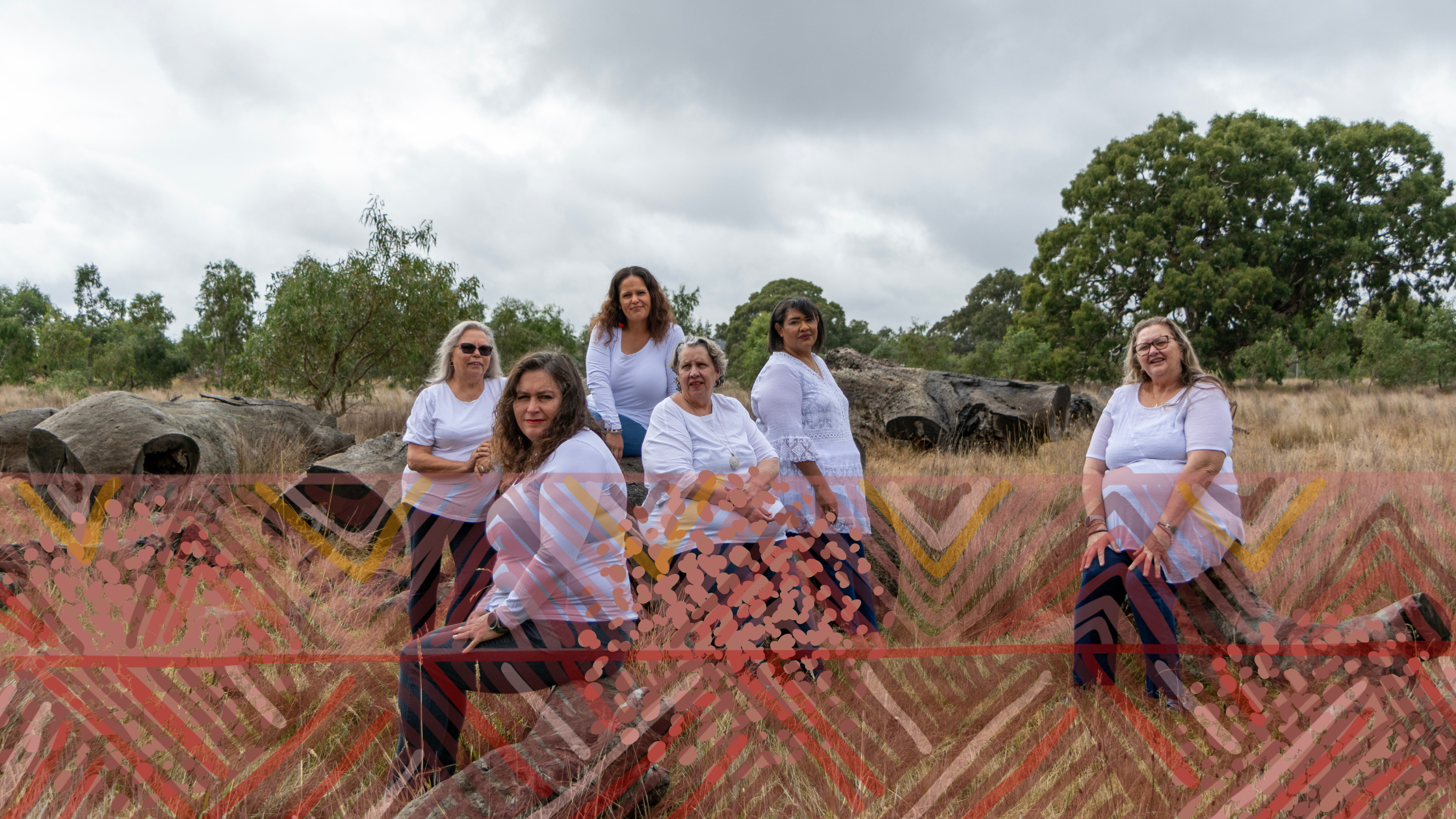 Group of six women standing and sitting outdoors in a grassy area with rocks and trees, wearing white tops and patterned skirts, under cloudy skies.