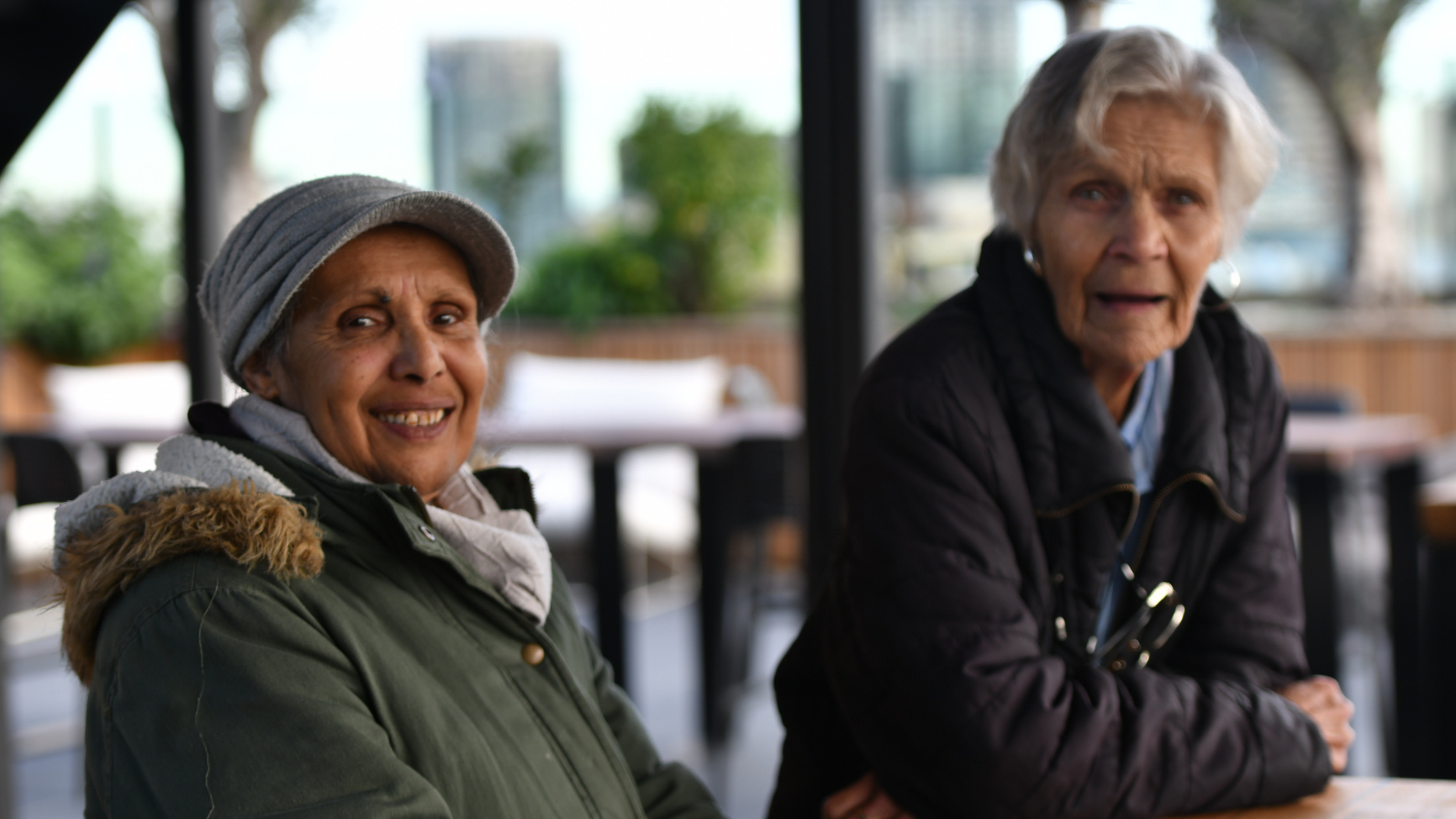 Two elderly women sitting at a table outdoors, engaged in conversation, with trees and a building in the background.