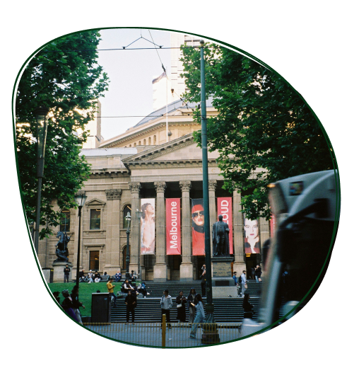 View of the Melbourne Museum with large banners on columns, surrounded by trees and people crossing the street in Melbourne, Australia.
