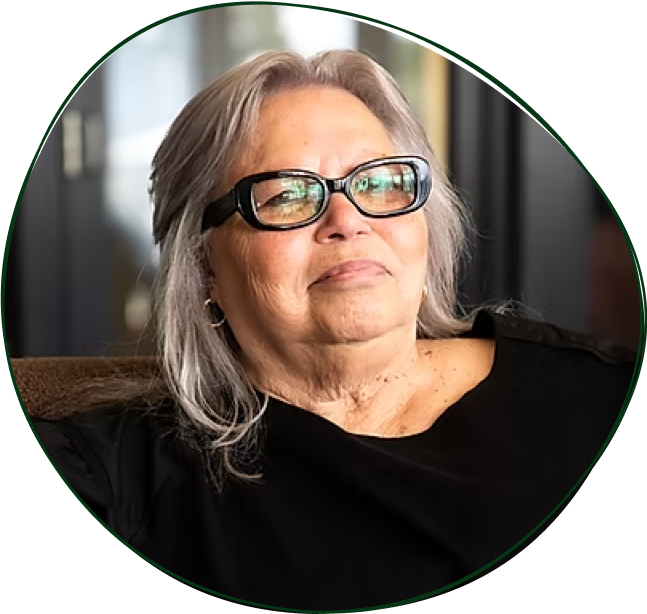 A woman with gray hair wearing large black glasses, sitting on a brown couch, looking confidently at the camera with a slight smile, indoors with a blurred background.