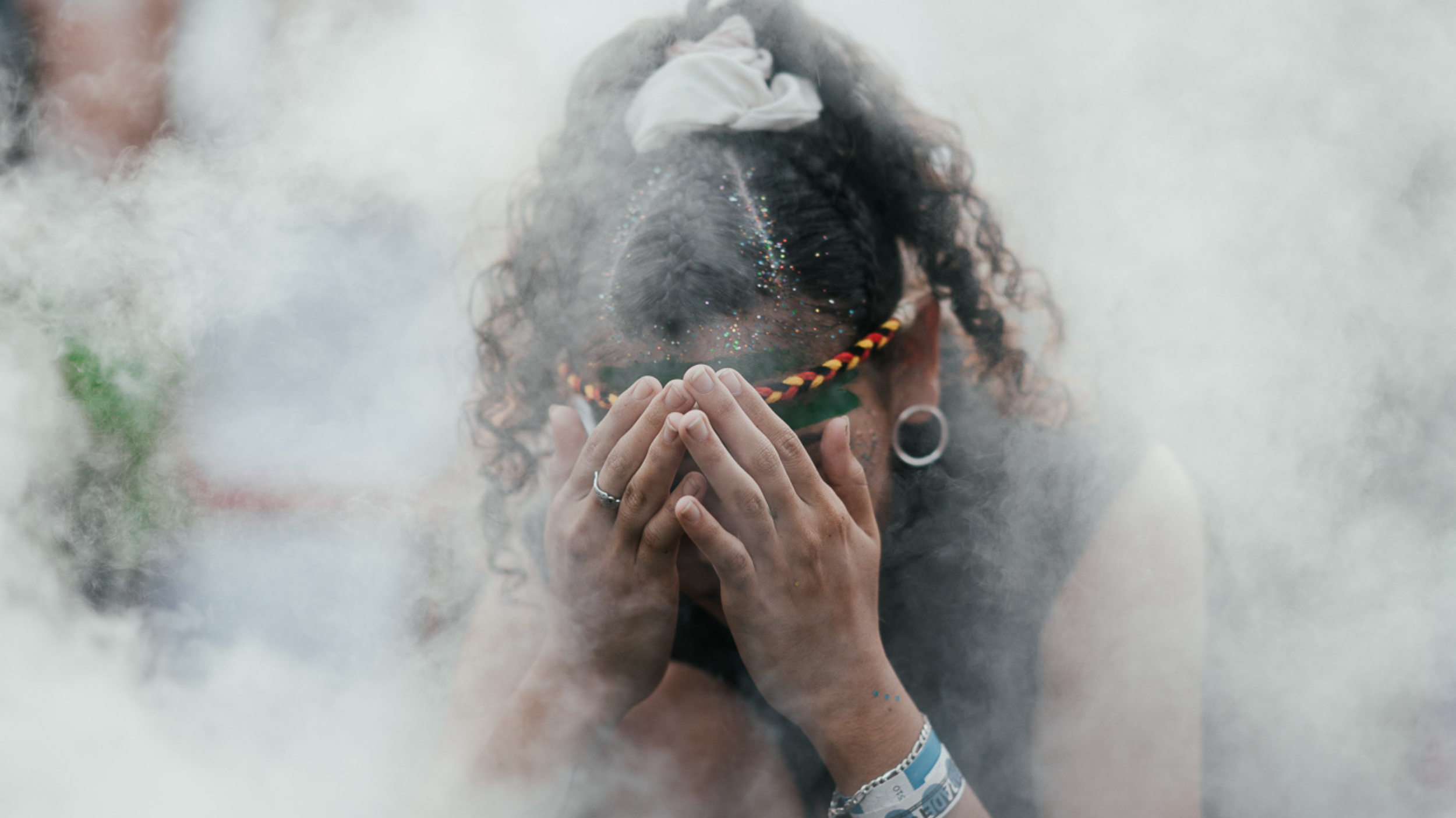 Person with curly hair and glitter on face, holding a smoking incense stick in front of face with eyes closed, in a smoky environment.
