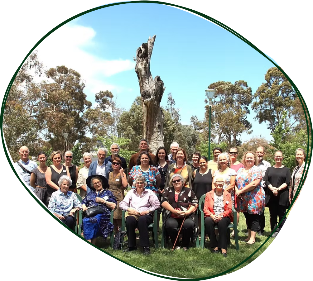 Group of people posing outdoors in front of a large tree with some people sitting and others standing, bright sunny day with clear blue sky.