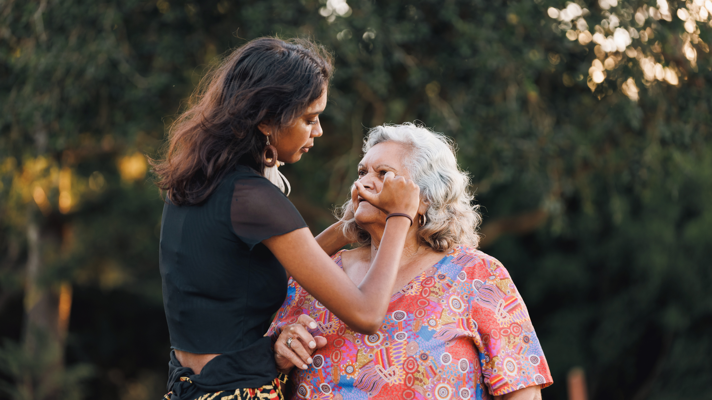 A young woman with dark hair and earrings gently holds a senior woman's face with both hands, in an outdoor setting with blurred green trees in the background.