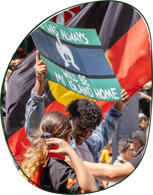 People protesting, holding a sign that says 'I was always a land, my will be island home,' while others hold flags in the background.