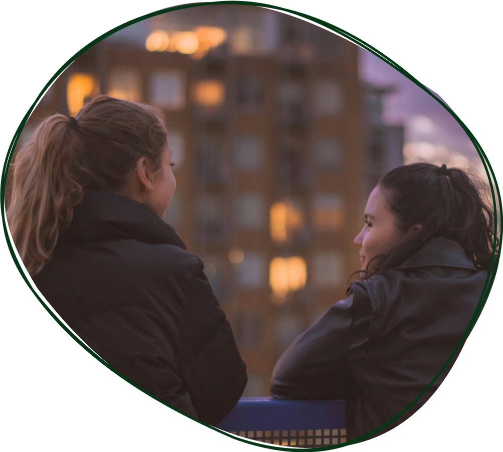 Two women chatting outdoors in an urban area at dusk, wearing black jackets, with a city building blurred in the background.