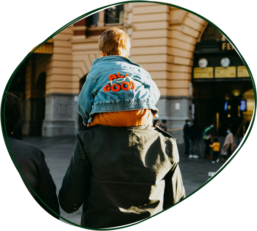 A child sitting on an adult's shoulders in front of a historic building, wearing a denim jacket with red letters that say 'ALL GOOD' on the back.