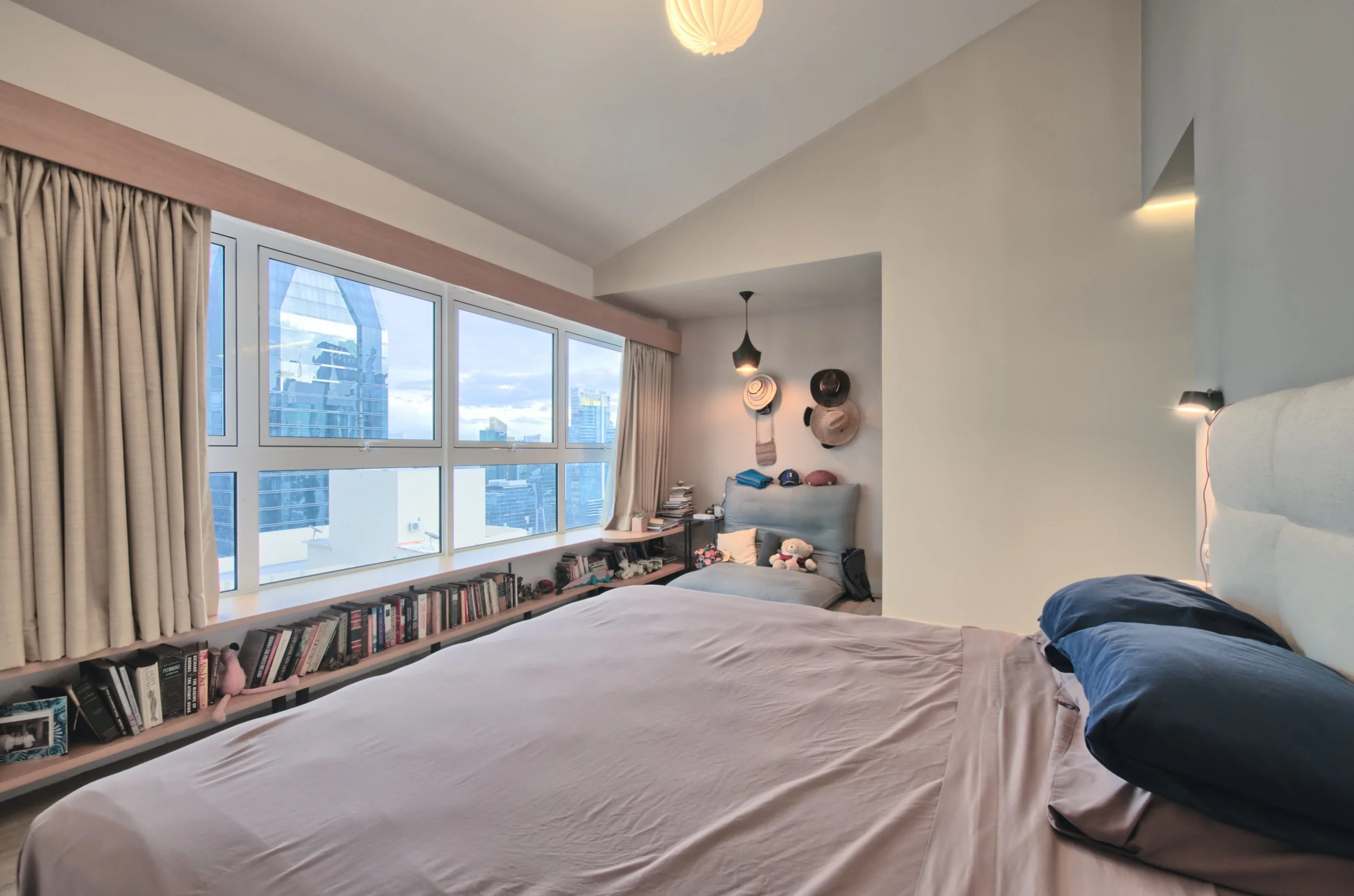 A bedroom with large windows, a bed with navy and beige bedding, a bookshelf along the window sill, and a sitting area with hats on the wall, stuffed animals, and books.