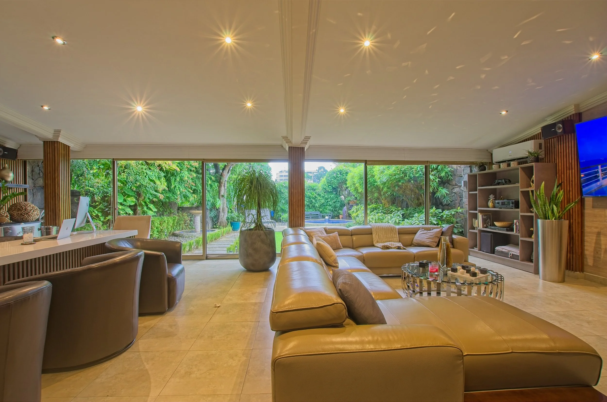 Living room with large windows showing lush greenery outside, beige sectional sofa, brown leather chairs, potted plants, media shelves, and a mounted TV.