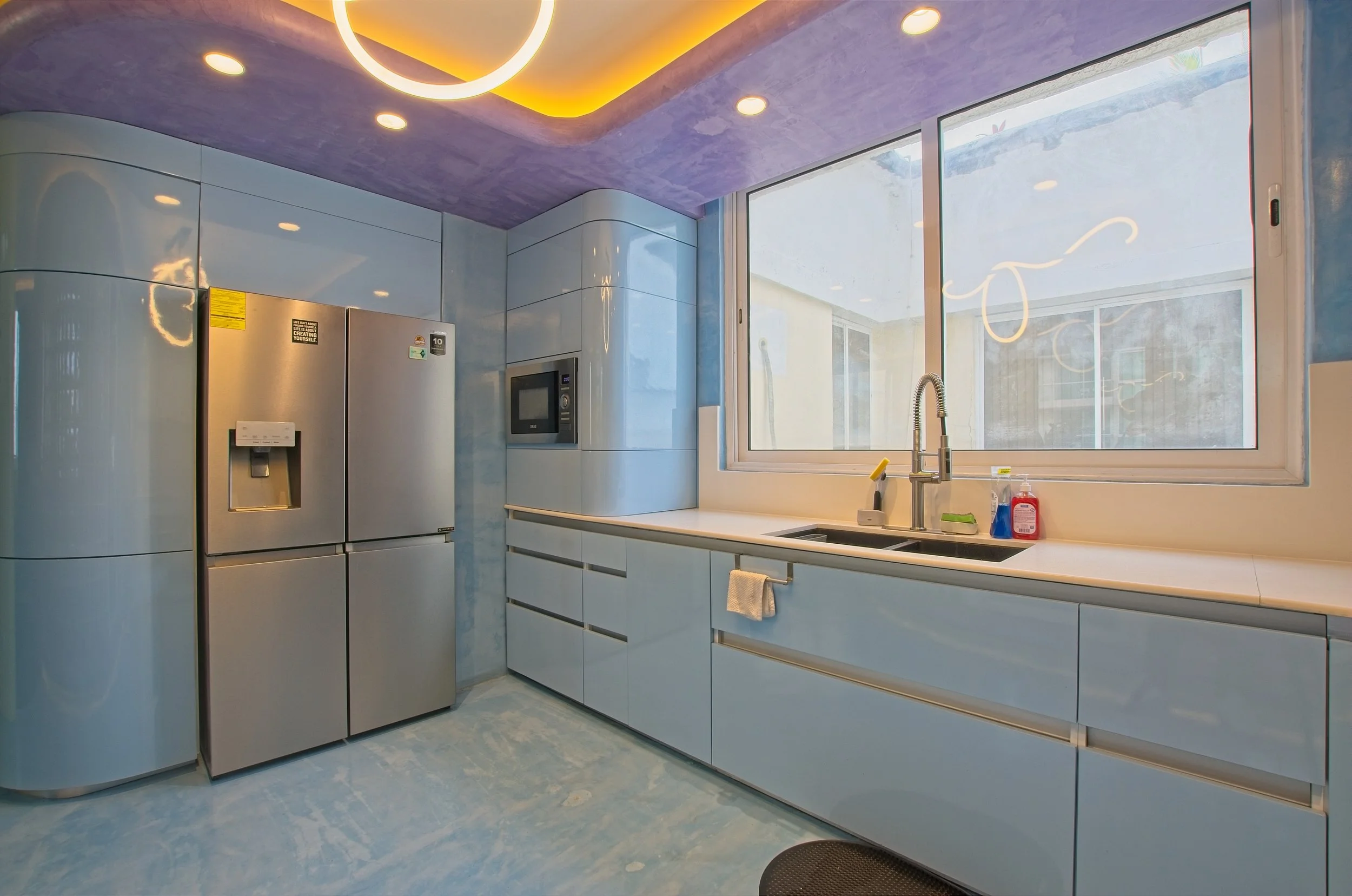 Cocina moderna con gabinetes azules brillantes, refrigerador de acero inoxidable, microondas y una ventana grande sobre el fregadero. El techo está pintado de morado, con luces empotradas y una lámpara de techo circular.