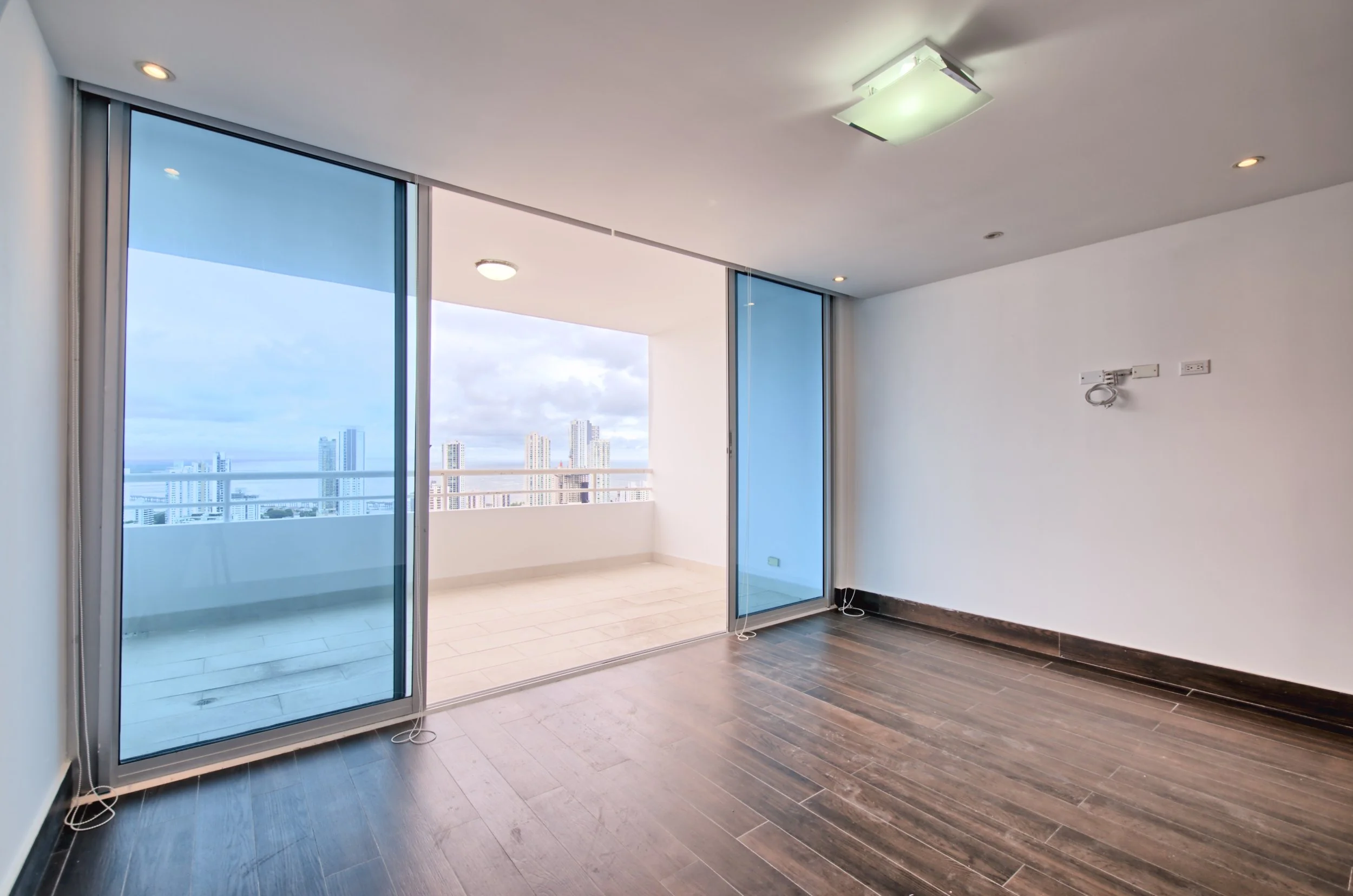 Empty room with wood floor, sliding glass door leading to a balcony, and city skyline view in the background.
