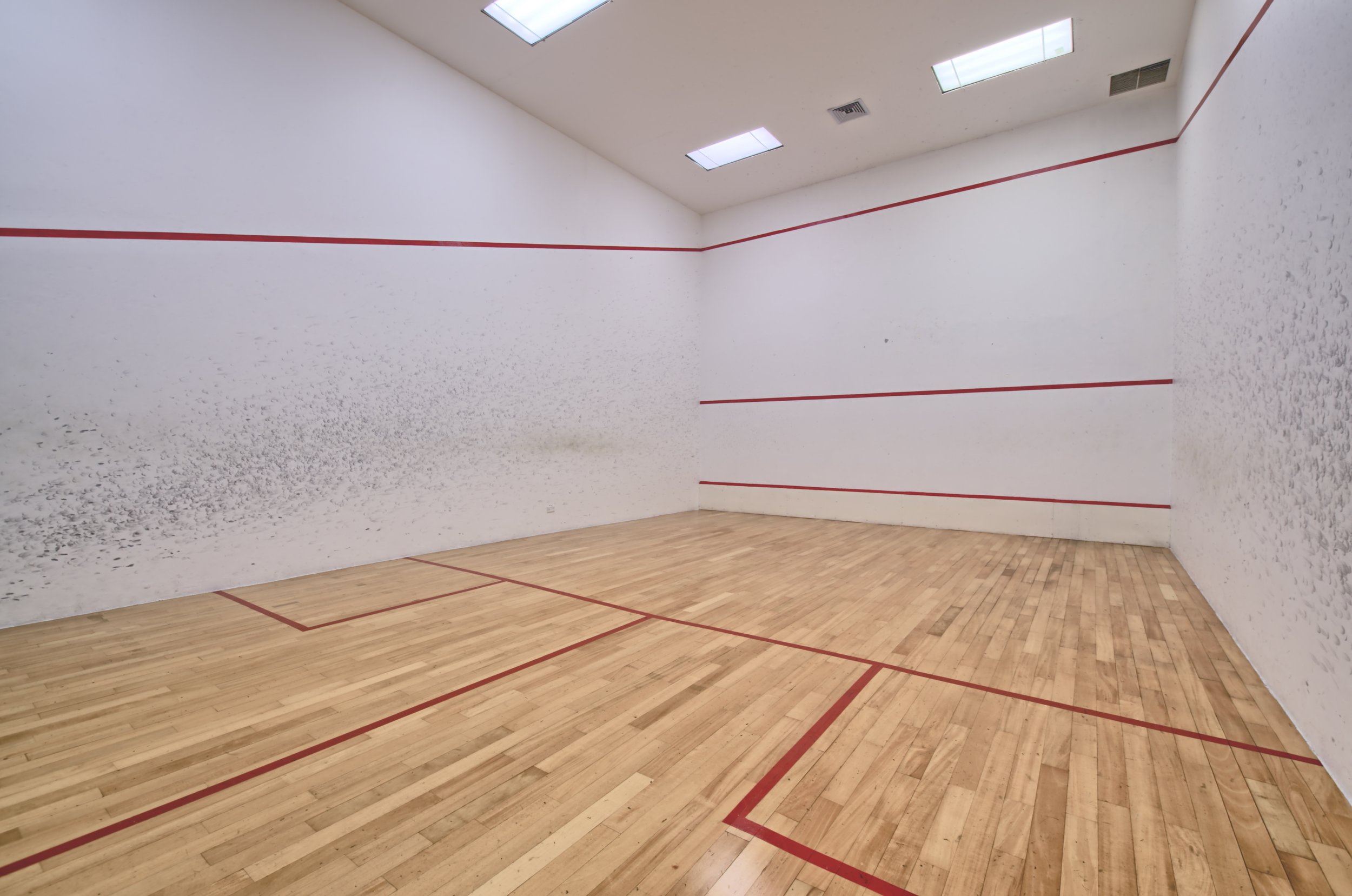 An indoor squash or racquetball court with white walls, red boundary lines, a wooden floor, and overhead lighting.