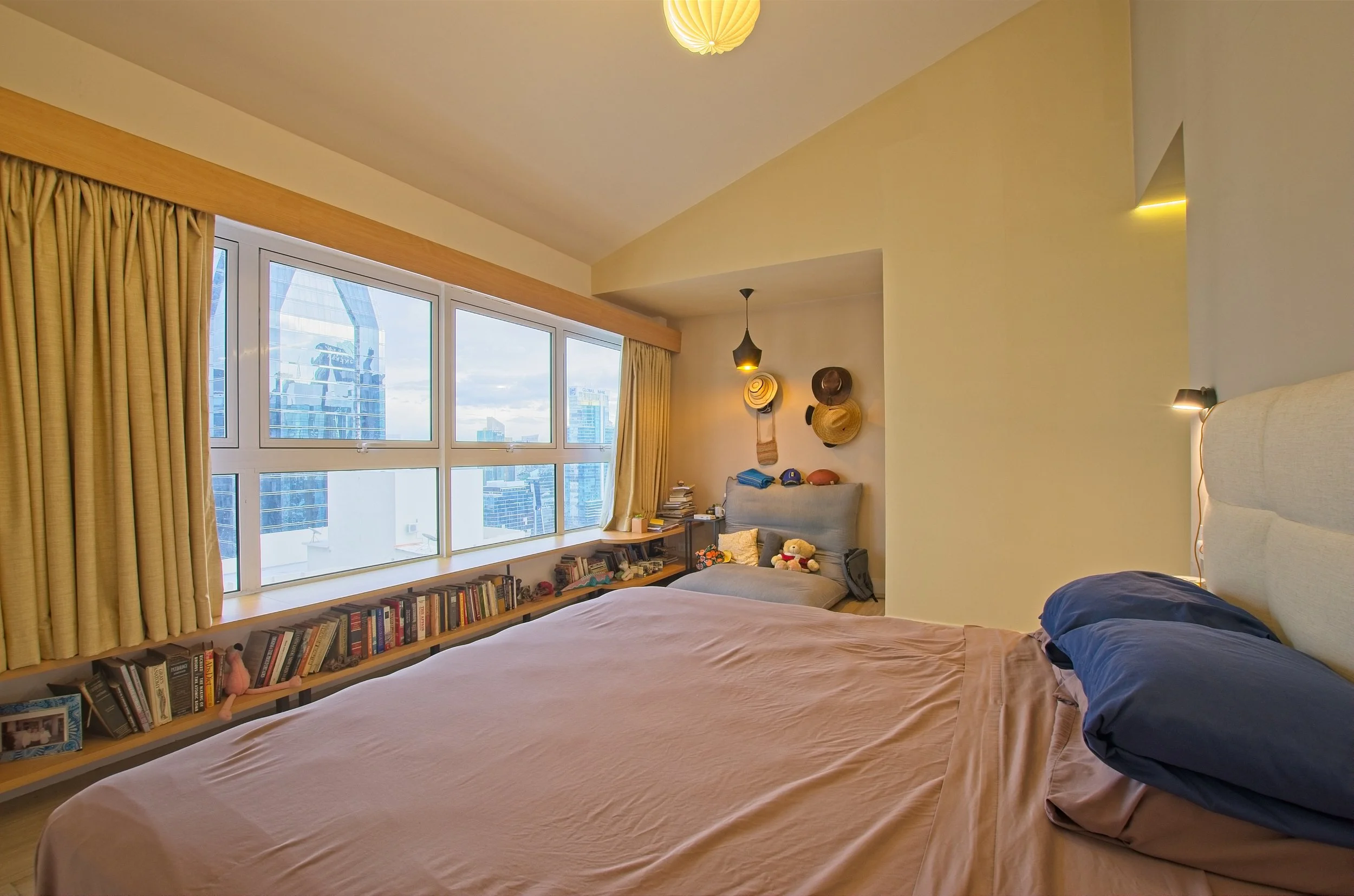 A bedroom with a large window showing a cityscape, a bed with pink sheets and blue pillows, a bookcase beneath the window, and a small seating area with hats and stuffed toys.