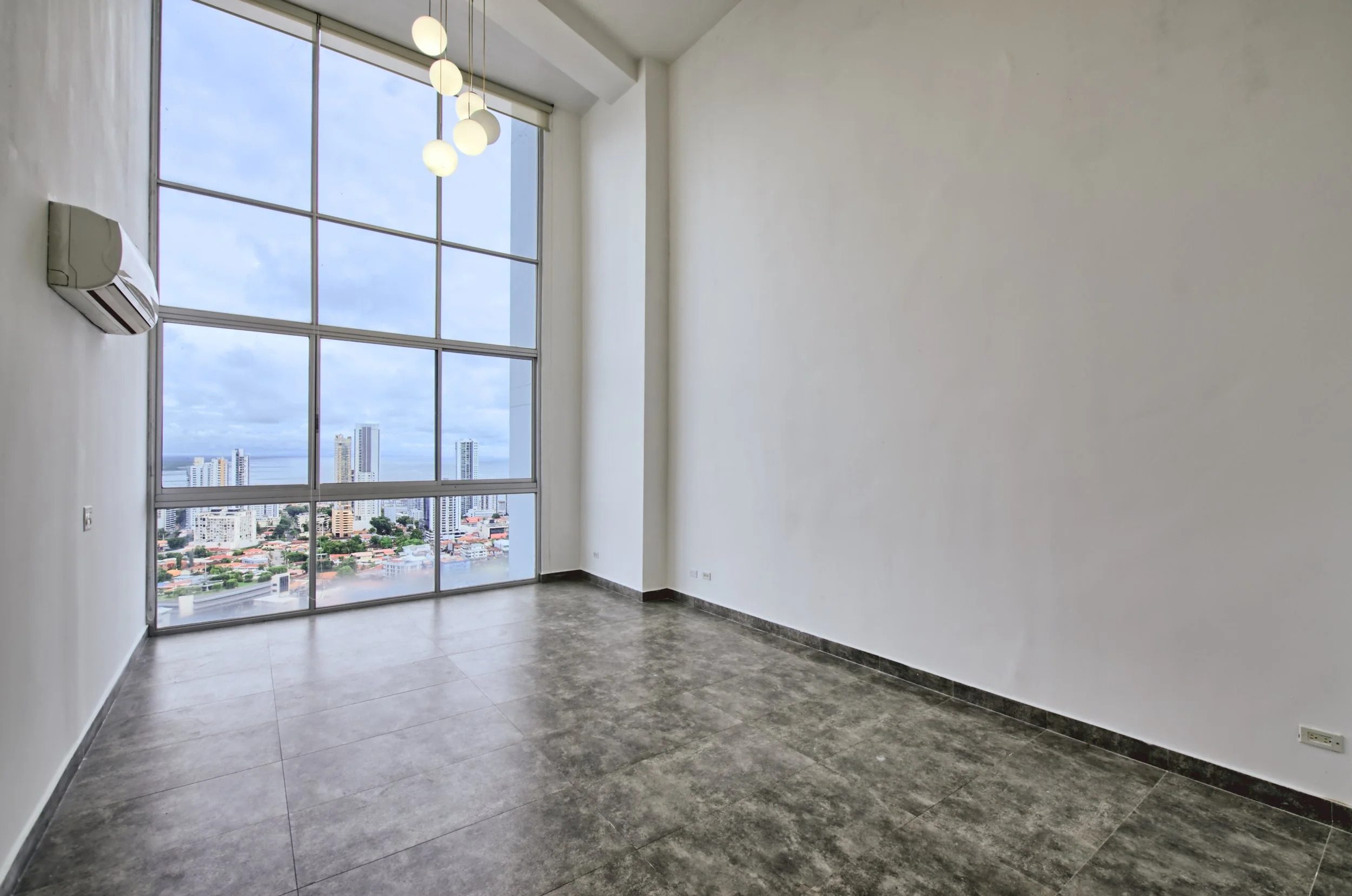 Empty room with large floor-to-ceiling window showing cityscape and ocean view, white walls, gray tile floor, wall-mounted air conditioning, modern hanging light fixture.