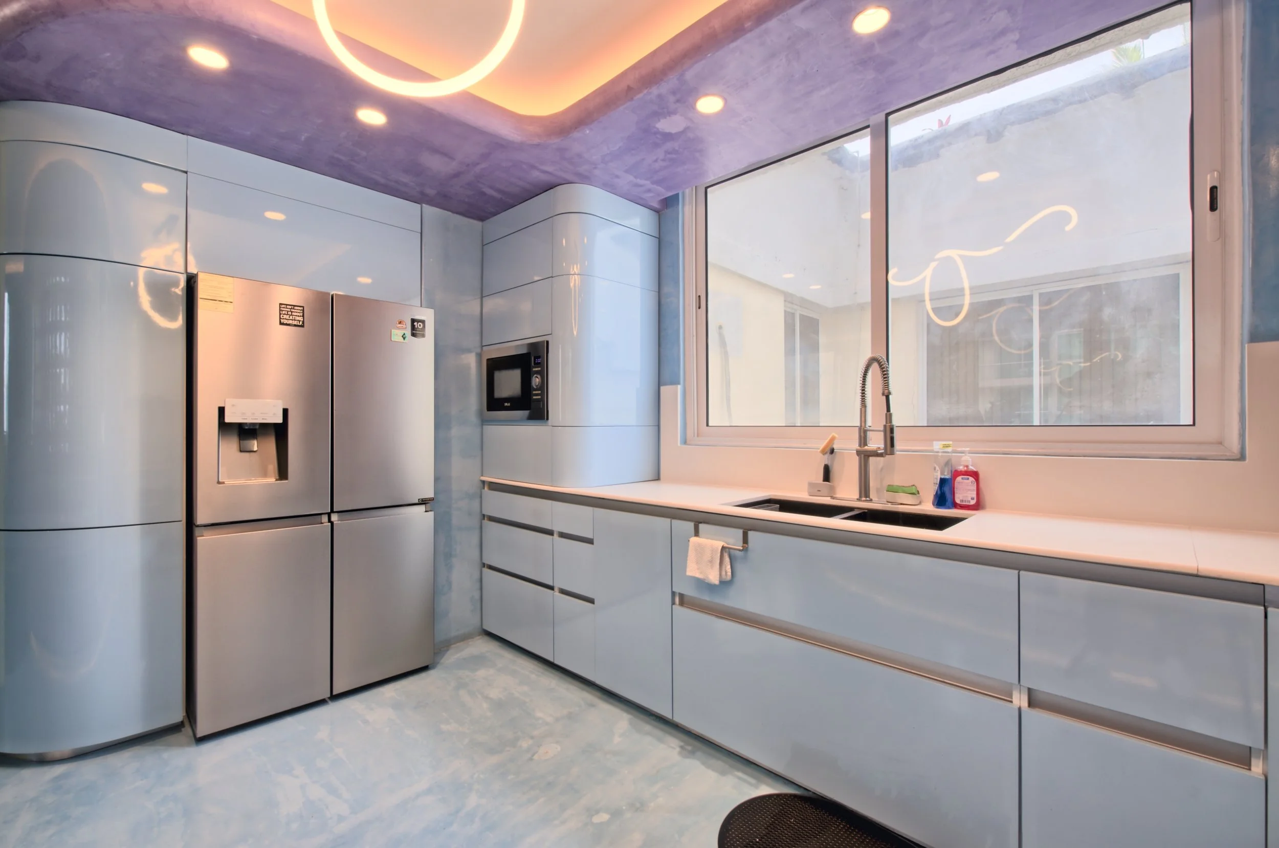 Modern kitchen with stainless steel refrigerator, white cabinetry, a microwave, and a large window above the sink with cleaning supplies on the countertop.