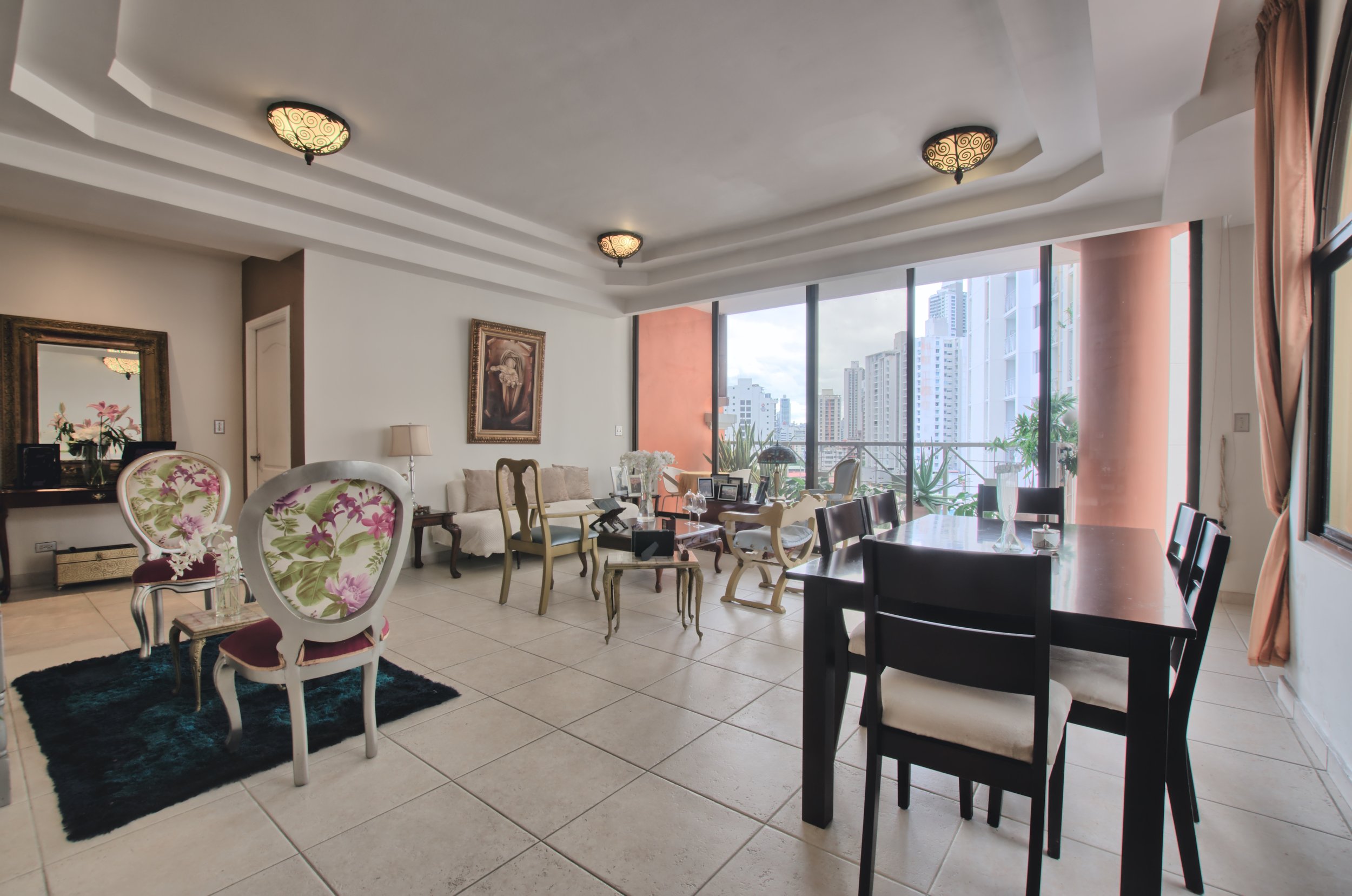 Spacious living and dining area with large floor-to-ceiling windows showing cityscape, floral-patterned chairs, white sofa, dining table, and decorative lighting.