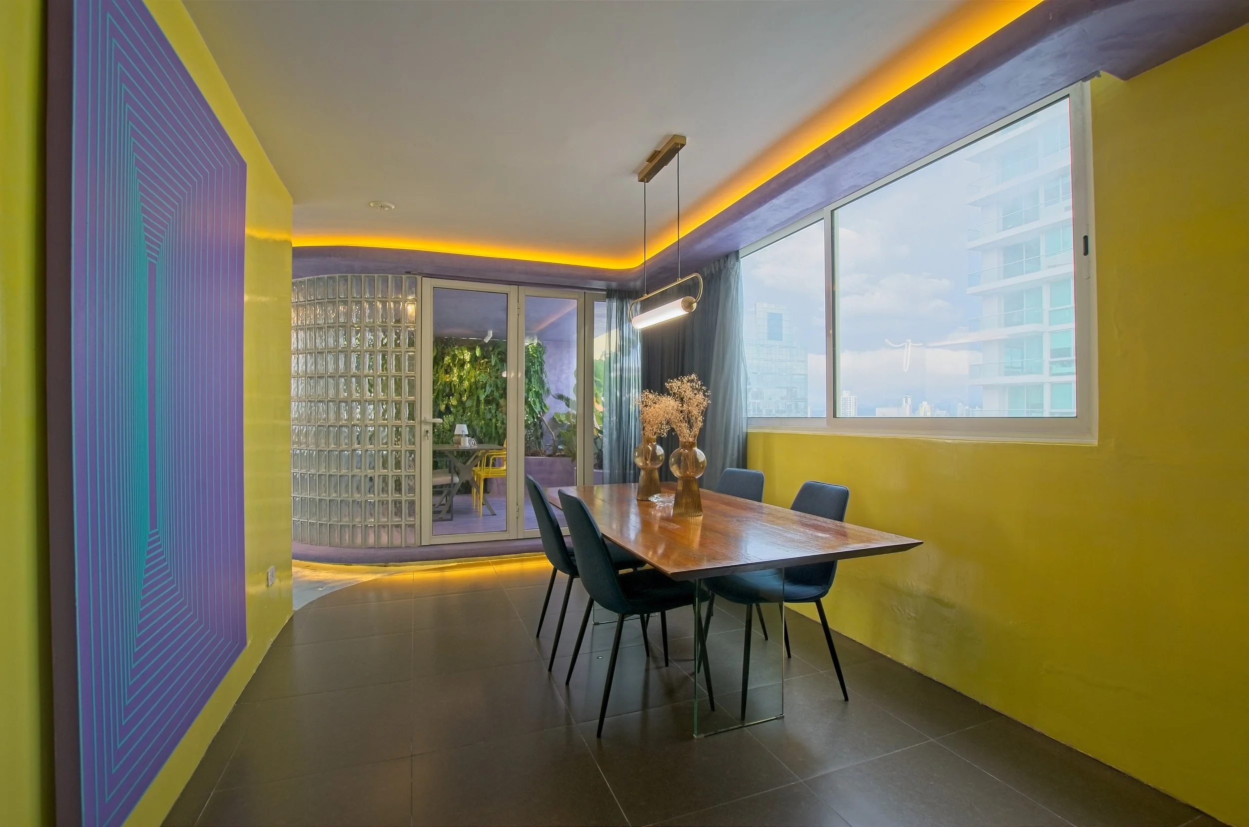 Comedor moderno con paredes amarillas, mesa de madera con cuatro sillas azules, dos jarrones con flores secas, grandes ventanas que muestran edificios de la ciudad y un balcón con vegetación visible a través de una pared de bloques de vidrio.