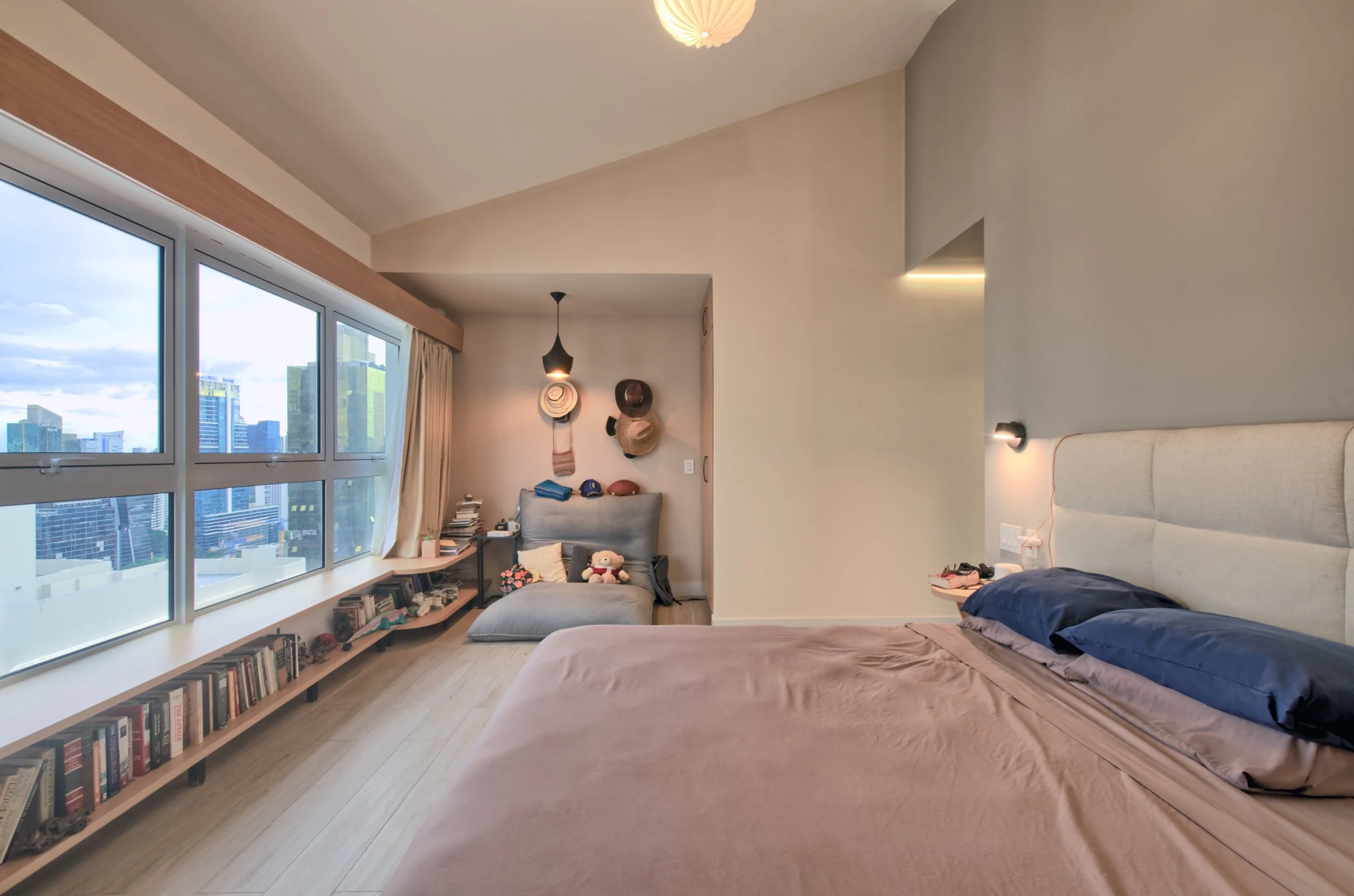 Modern bedroom with large window overlooking city skyline, bed with beige headboard, blue pillows, bedside lights, and a small sitting area with books, bookshelves, and hats on wall.