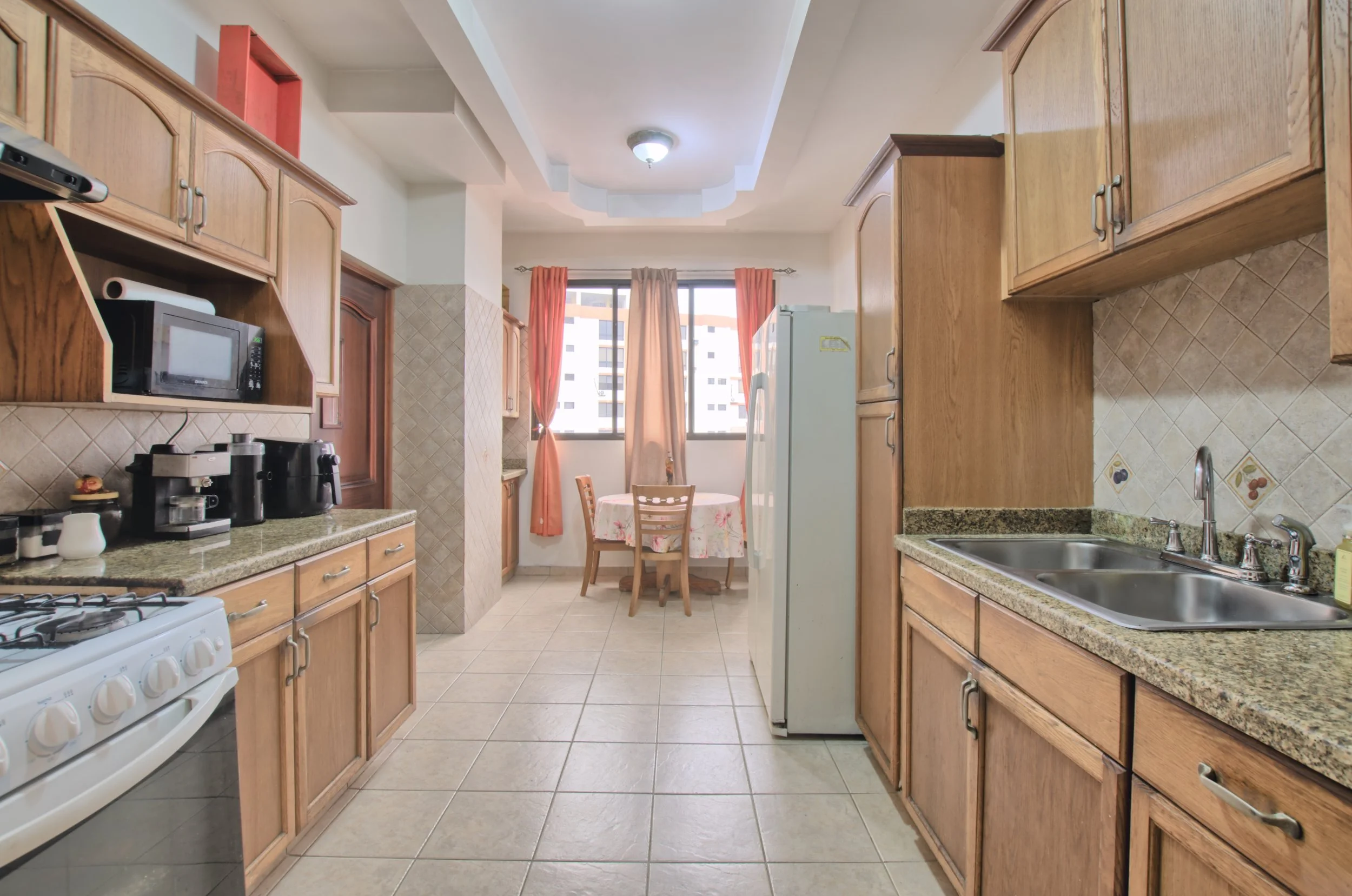 Kitchen with wooden cabinets, granite countertops, a stove, microwave, coffee maker, double sink, refrigerator, and a circular dining table with chairs near large windows with peach curtains.