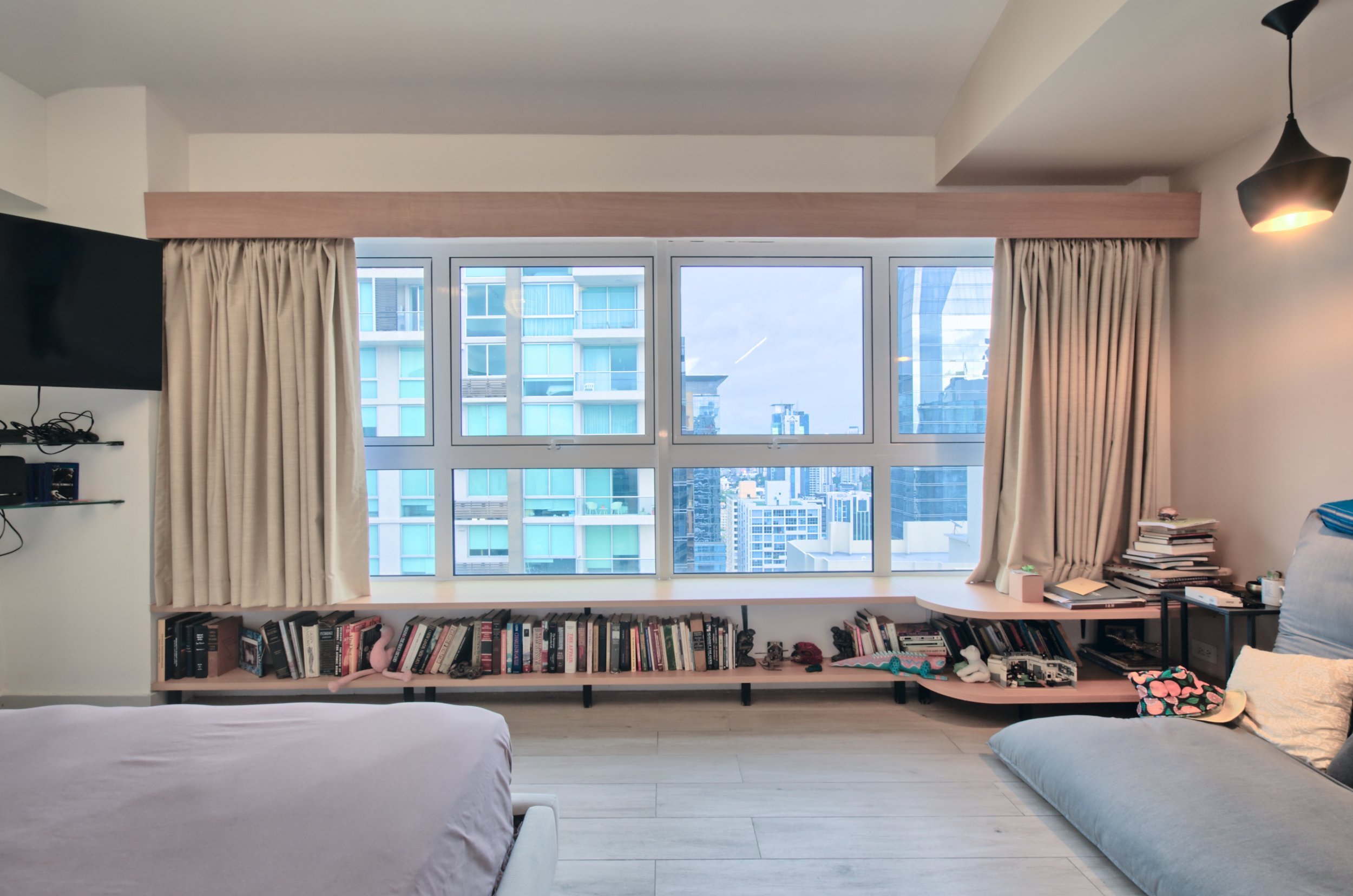 Living room with large window overlooking city buildings, beige curtains, bookshelves under window, bed in foreground, gray sofa with pillow and blanket on right side, small side table with stacks of books and miscellaneous items, wall-mounted TV on left.