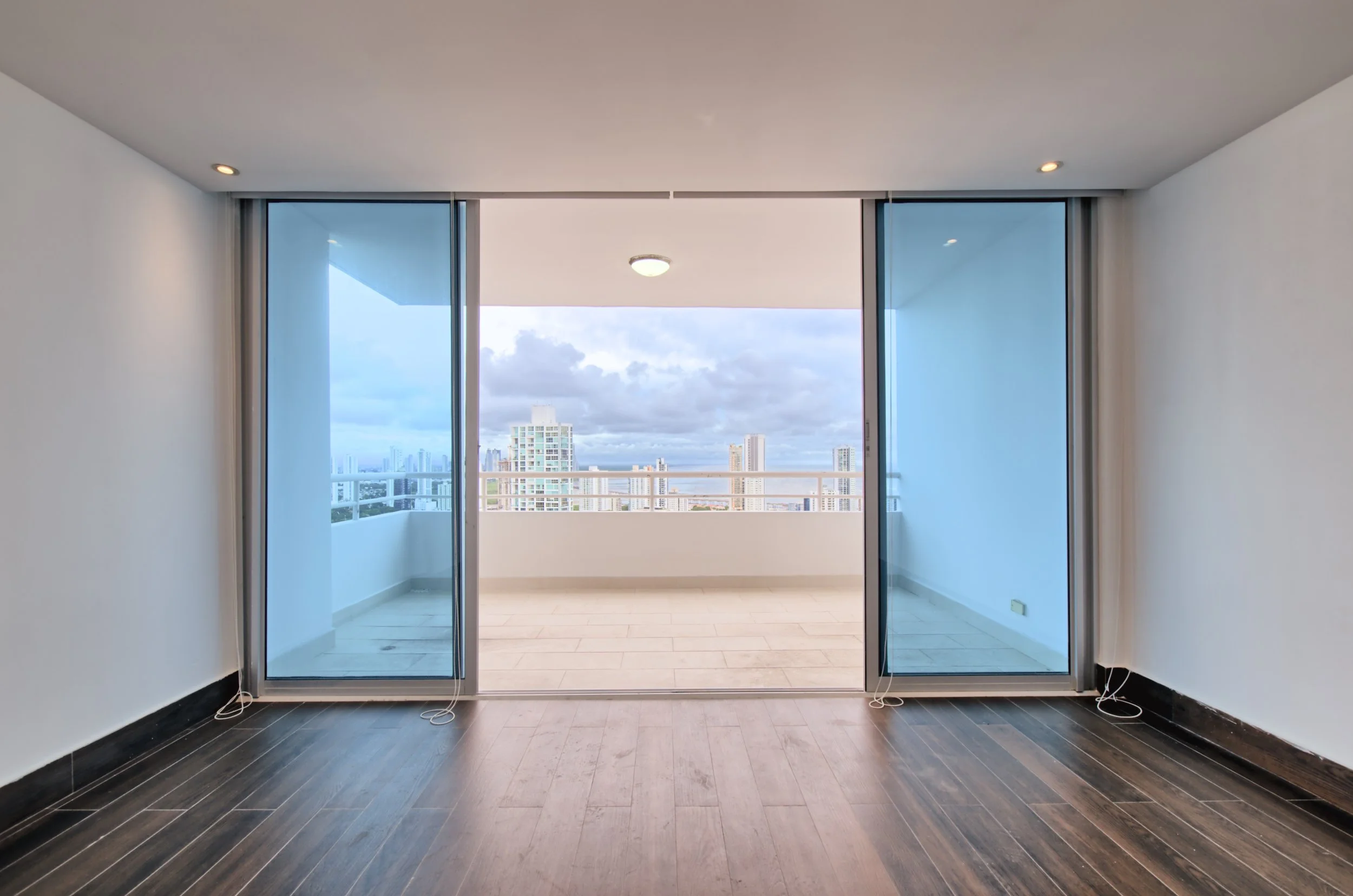 Empty living room with wood flooring and large sliding glass doors leading to a balcony with city skyscraper view.