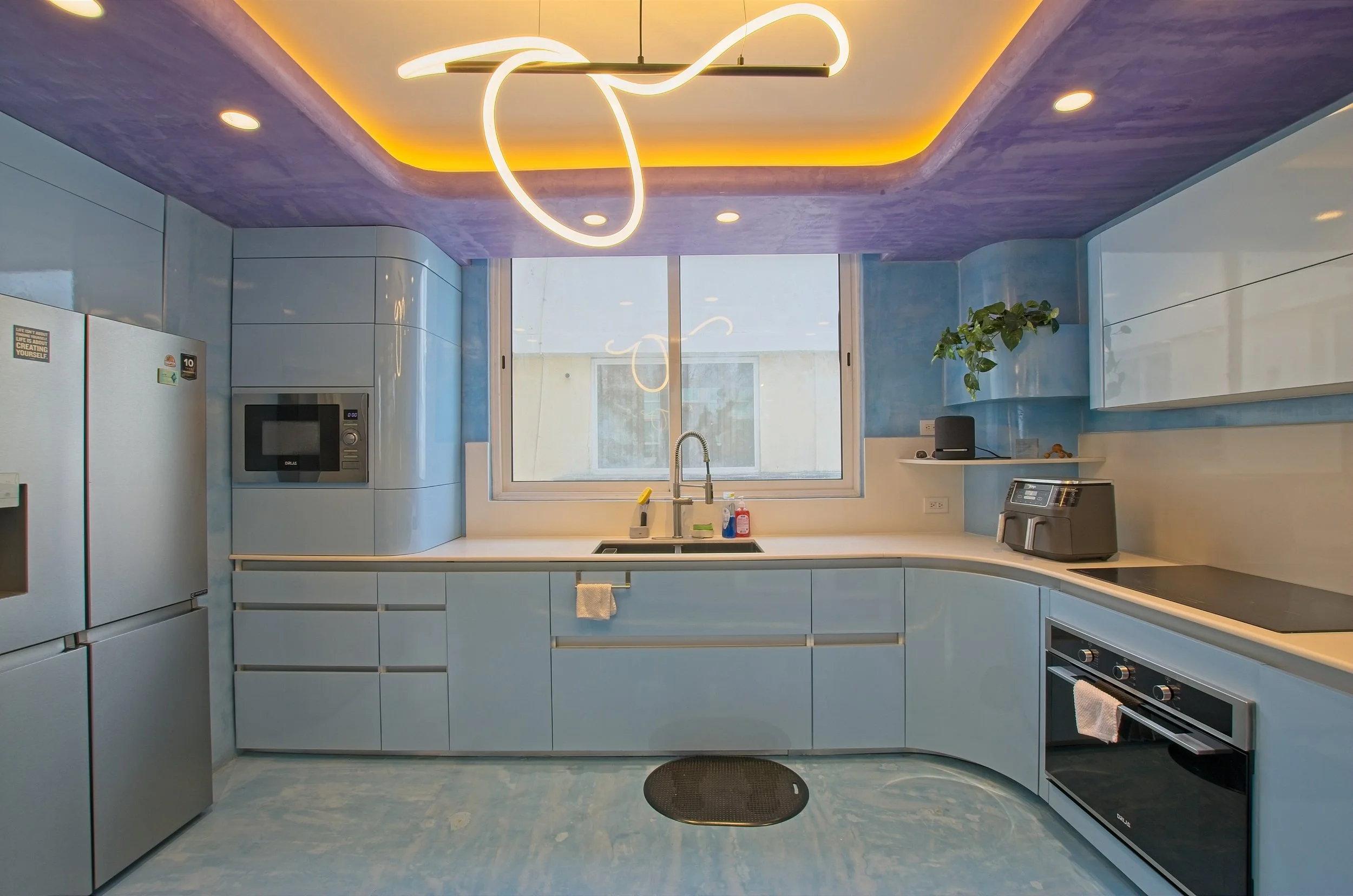Modern kitchen with blue cabinets, white countertops, and a large window. Features include a refrigerator, microwave, toaster, and stove, with decorative ceiling lighting and a potted plant.