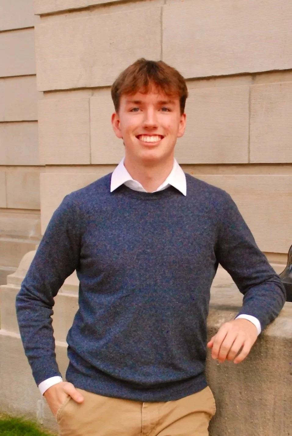 Young man with brown hair smiling and wearing a dark sweater over a collared shirt, beige pants, standing outdoors near a stone building with steps and a black lamppost.