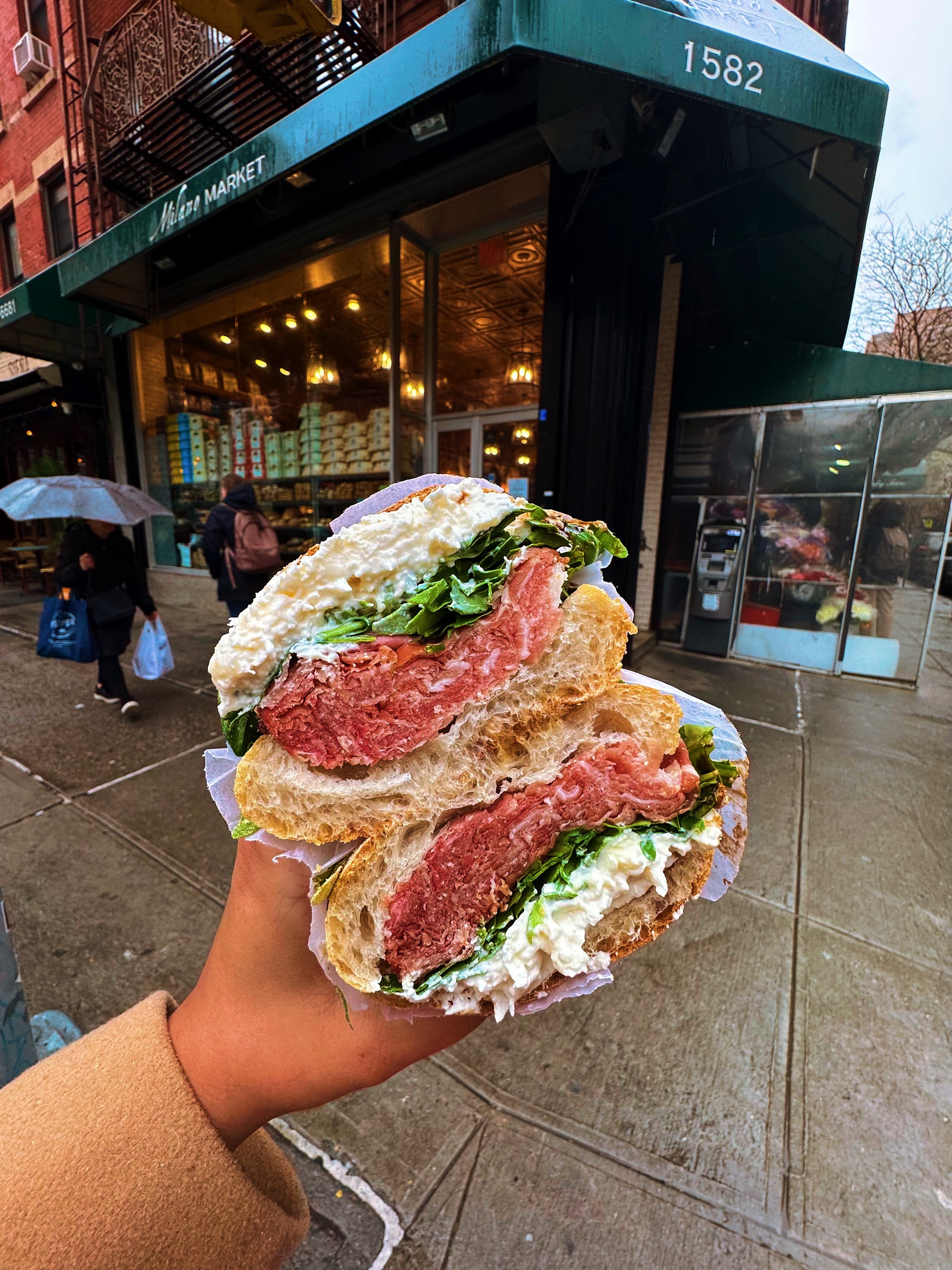 Close-up of a hand holding a double-cut sandwich with steak, lettuce, and mayonnaise outside a bakery on a rainy day.