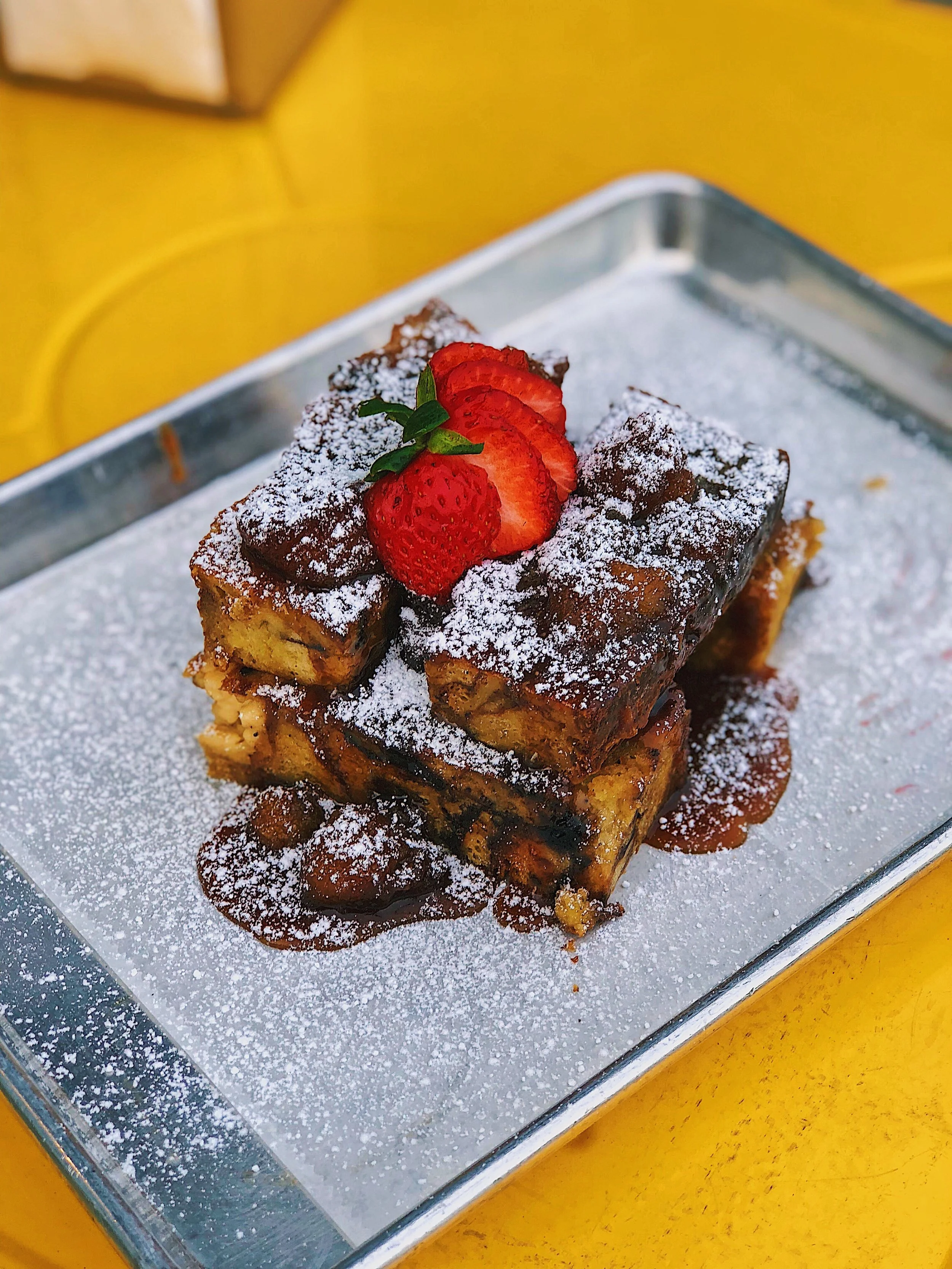 Stack of French toast topped with strawberries, powdered sugar, and syrup on a metal tray with yellow background.