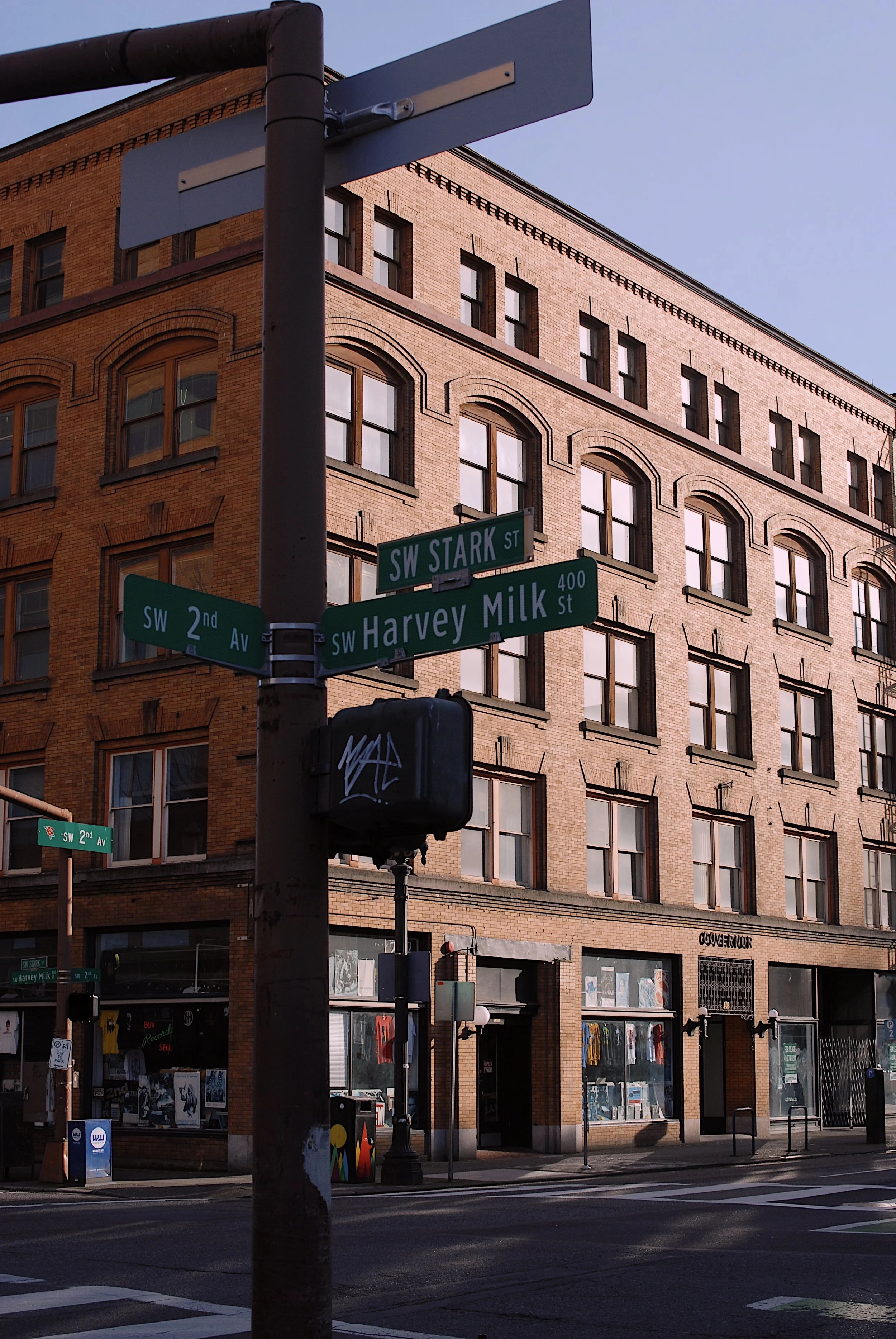 Street corner with street signs for SW Stark Street, SW Harvey Milk Street, and SW 2nd Avenue in front of a brick building with retail shops on the ground floor.